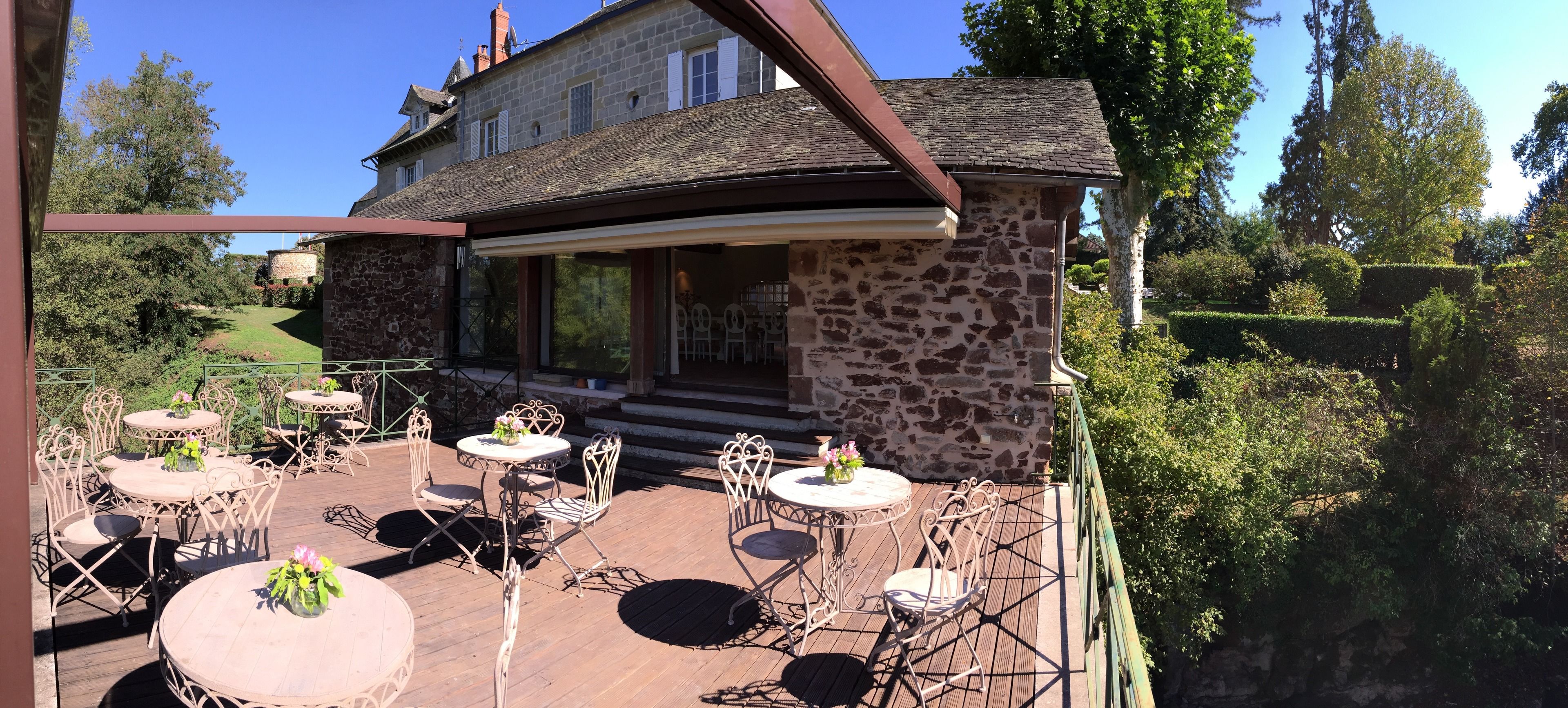 A stone building with a balcony, furnished with tables and chairs, surrounded by greenery.
