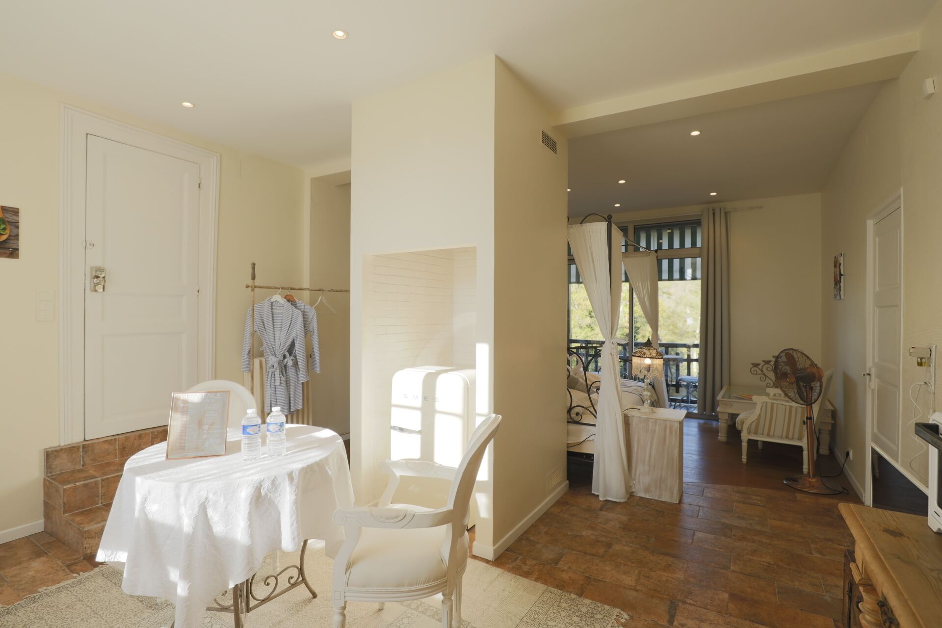 A spacious room with a white table, a chair, and a bed. There is a window with curtains, a fan, and a door.