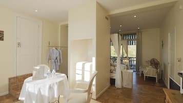 A spacious room with a white table, a chair, and a bed. There is a window with curtains, a fan, and a door.