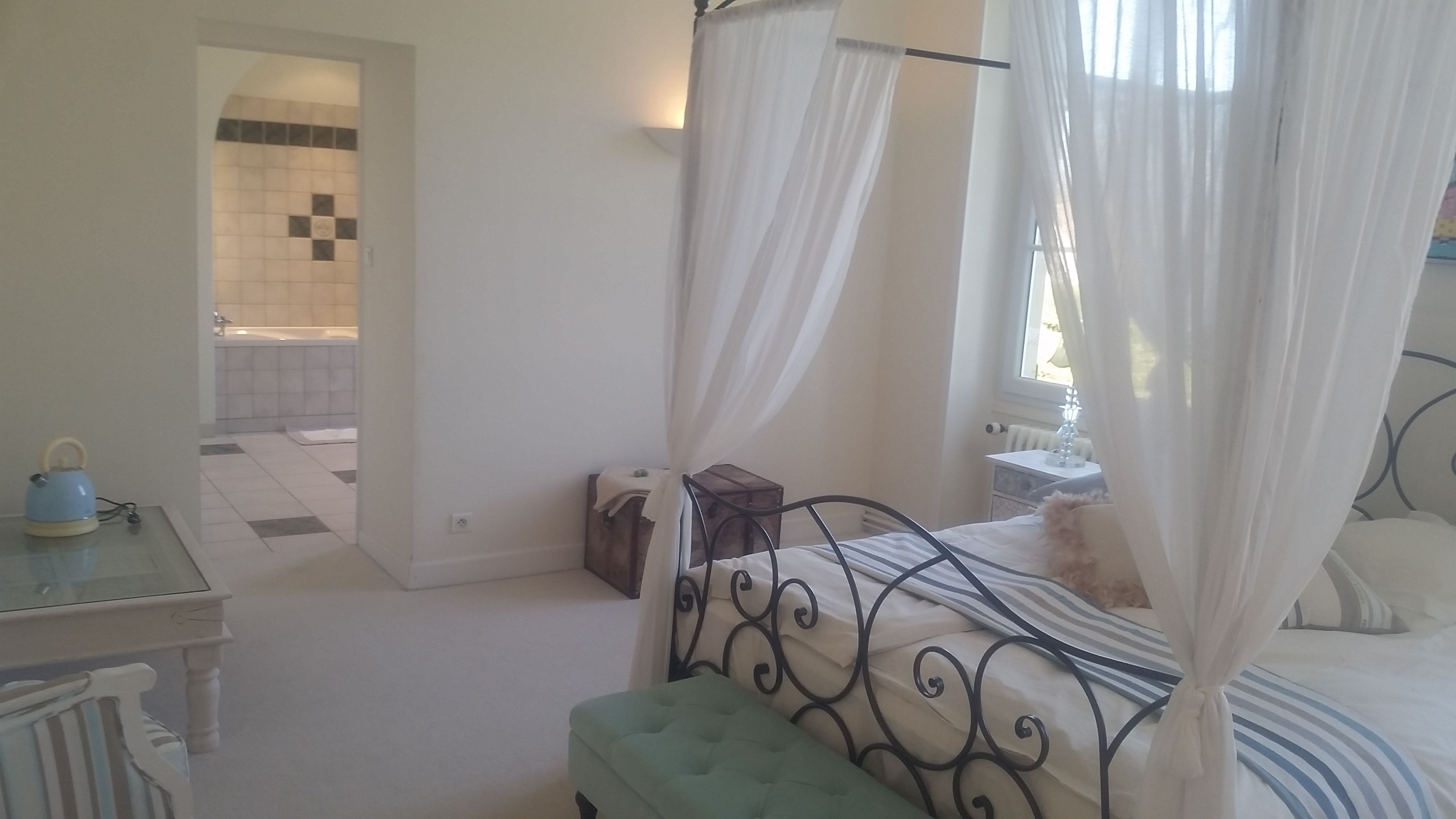 A bedroom with a bed, a bedside table, a window with curtains, and a bathroom visible through an open door.