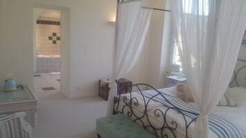 A bedroom with a bed, a bedside table, a window with curtains, and a bathroom visible through an open door.