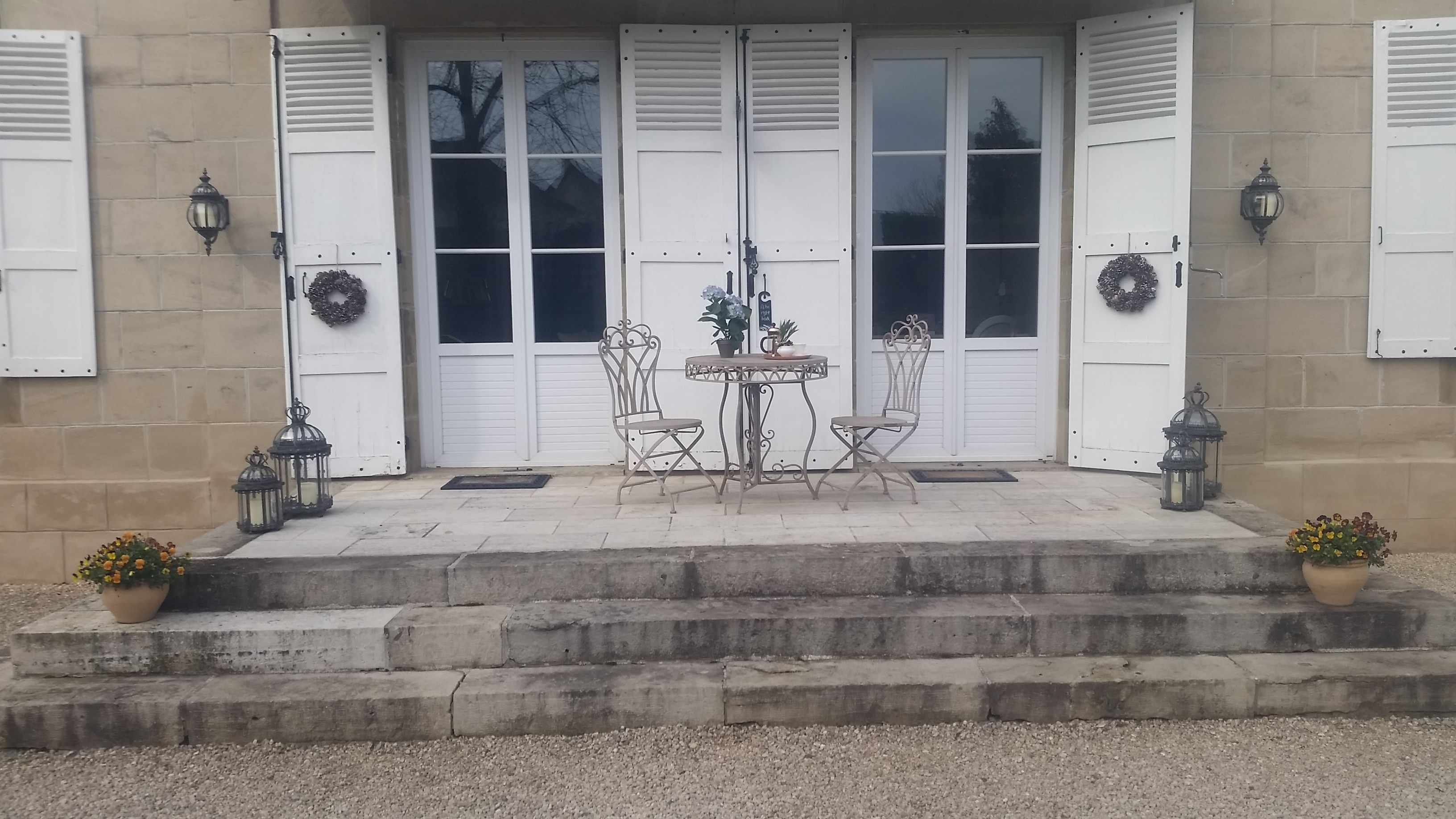 A building entrance with white shutters, a small round table with chairs, and two potted plants.