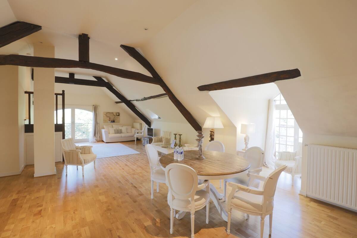 A spacious loft with high ceilings, exposed wooden beams, a central wooden dining table surrounded by white chairs, and a cozy seating area with a white sofa and armchairs.