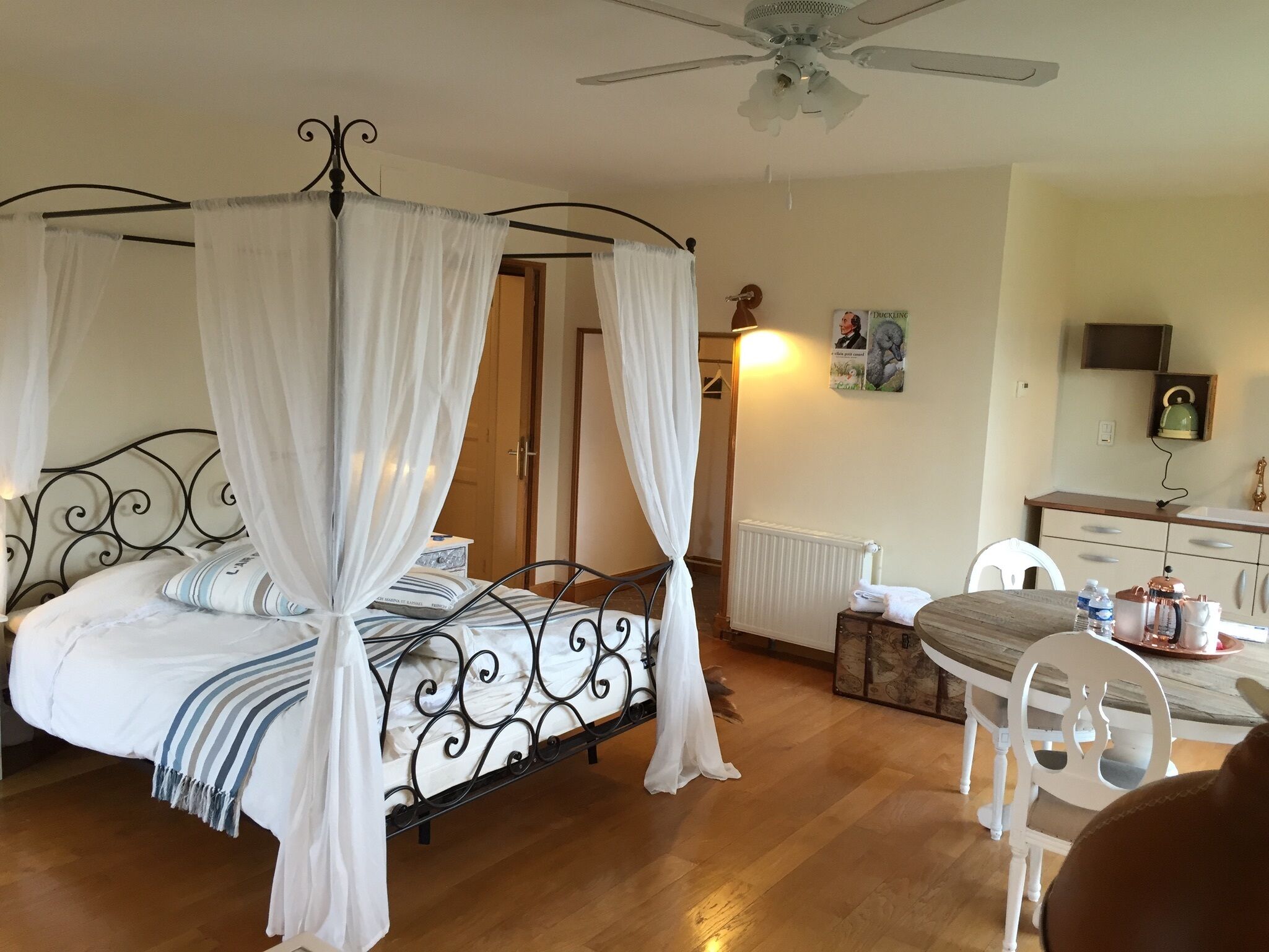A four-poster bed with white curtains, a wooden floor, a ceiling fan, a radiator, a small table with a chair, and a wall-mounted shelf with a kettle and a cup.