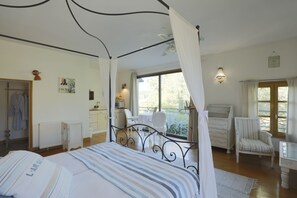 A bedroom with a four-poster bed, a sitting area with a table and chairs, a view of the garden through a window, and a bathroom with a shower and a towel rack. - Moulin Bleu de Vézère (Saint-Pantaleon-de-Larche)