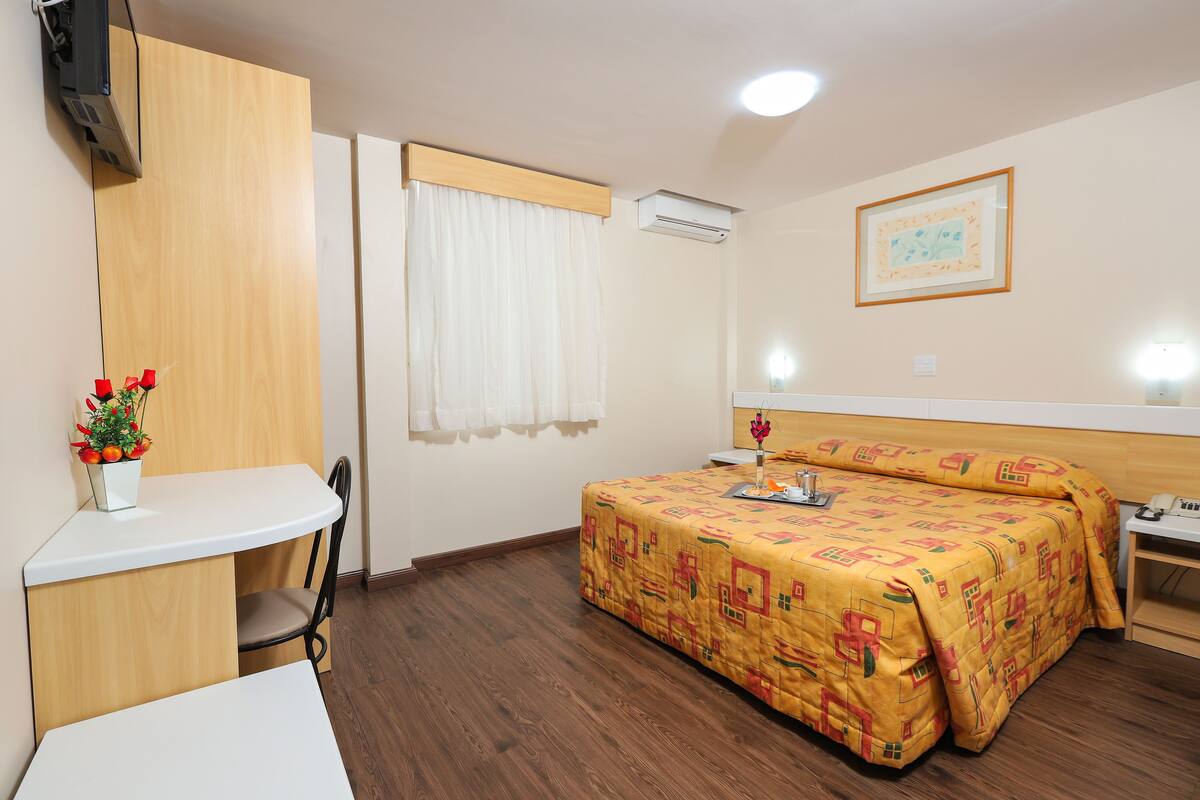 Ein Hotelzimmer mit Bett, Schreibtisch, Stuhl, einem kleinen Tisch, einer Topfpflanze und einem gerahmten Bild an der Wand.