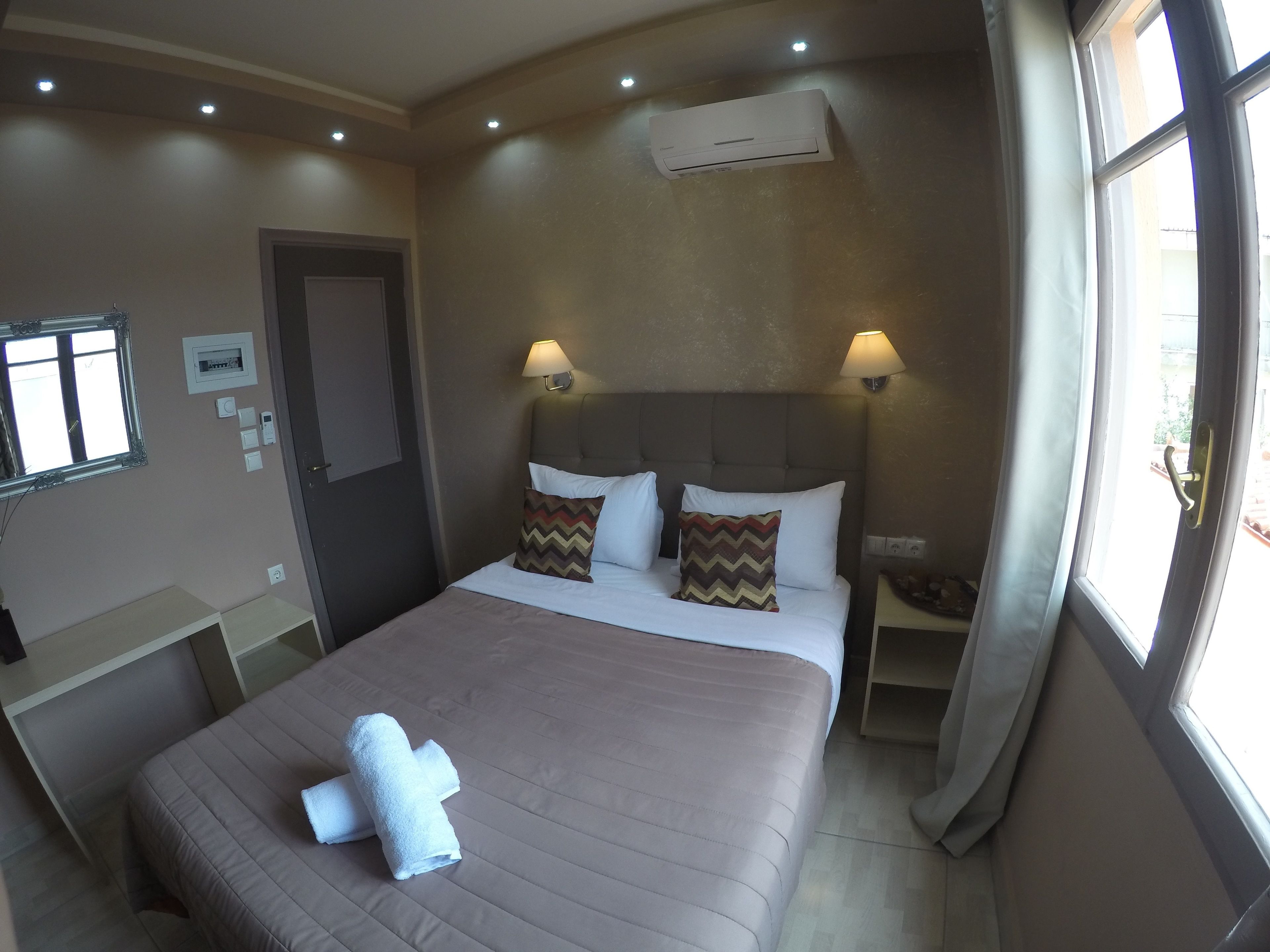 A hotel room with a bed, two bedside lamps, an air conditioning unit, a window with curtains, and a door.