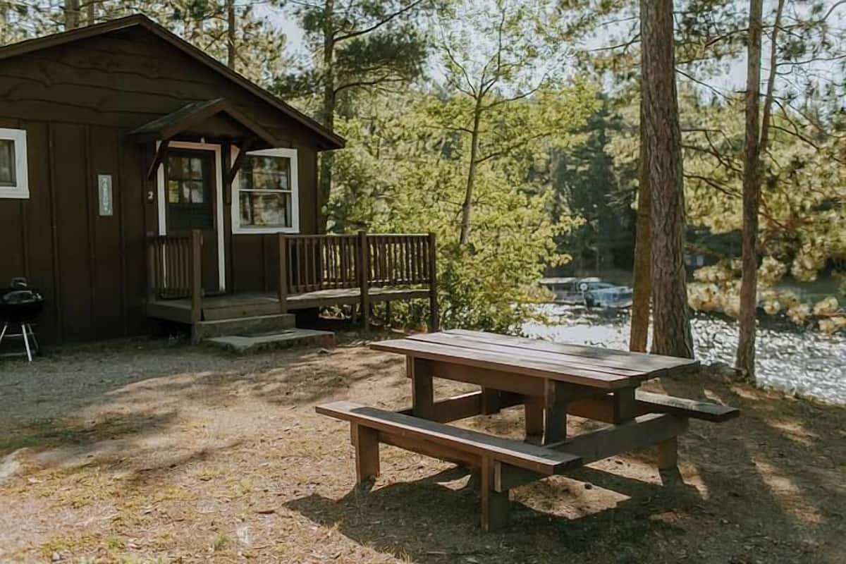 Una cabaña rústica con una mesa de picnic al frente, rodeada de árboles y una masa de agua.