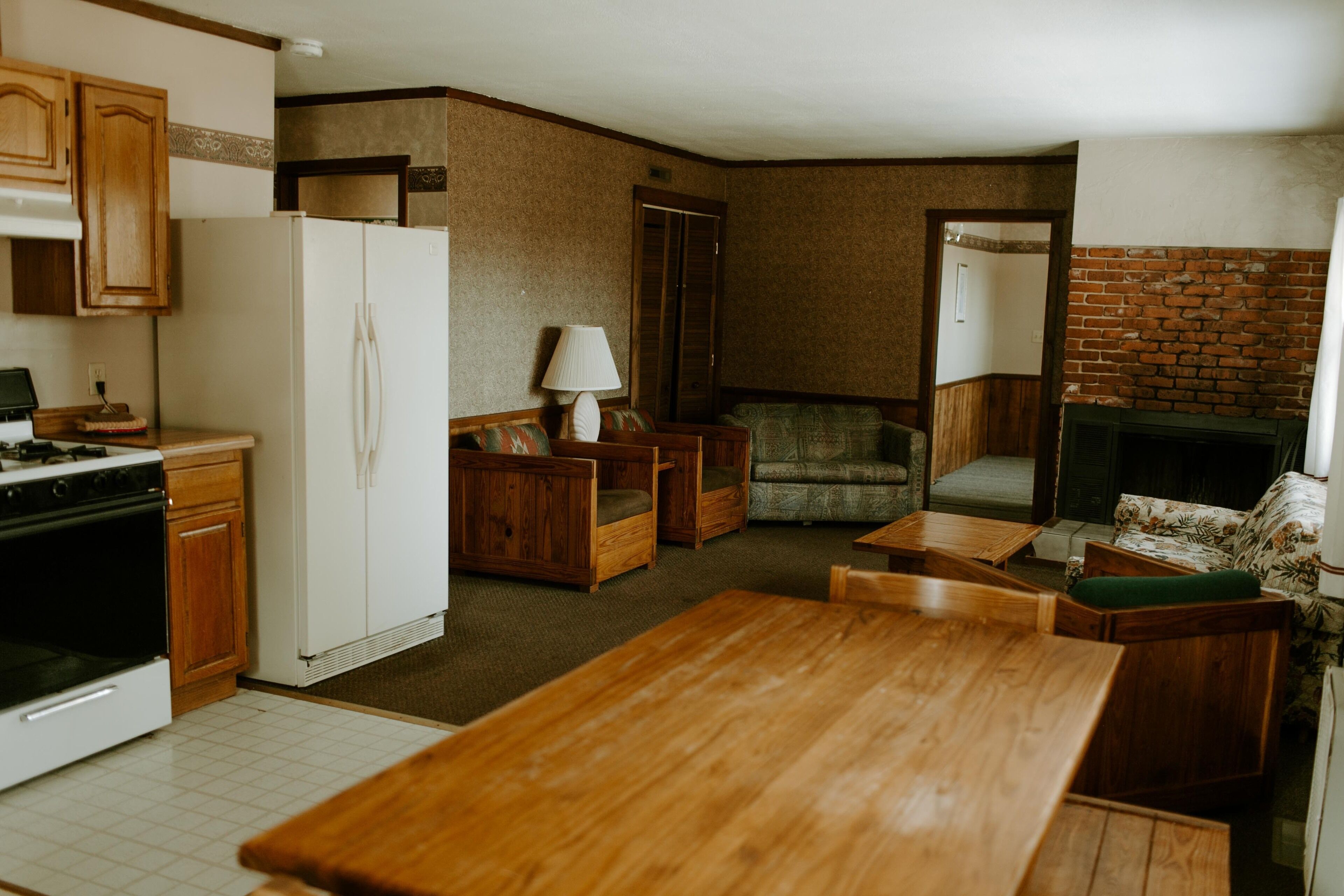 A compact kitchen with wooden cabinets, a refrigerator, and a stove. There is a dining area with a wooden table and chairs. A sofa and a chair are placed in the living room area.