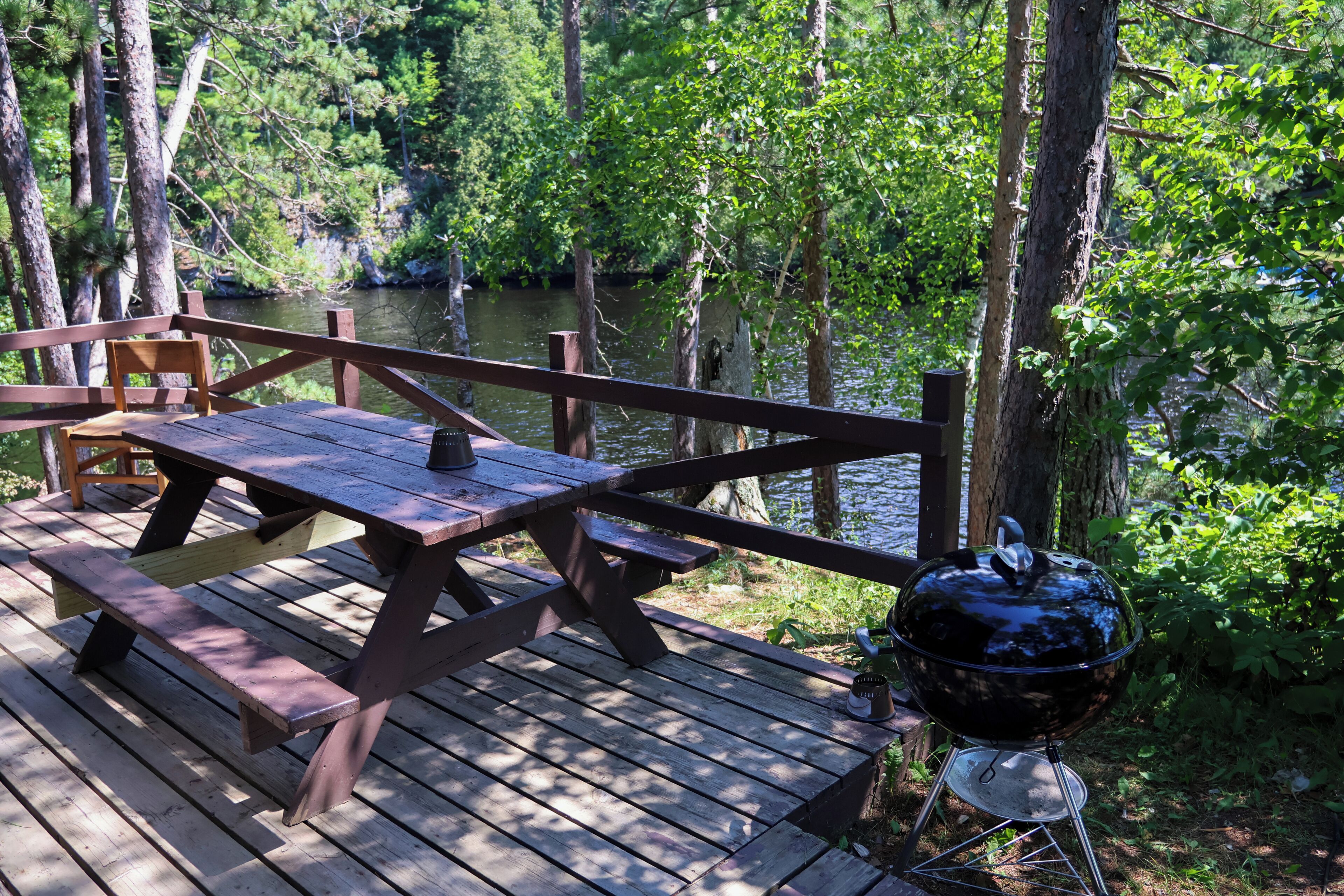 Een houten terras met picknicktafels en een barbecue, met uitzicht op een rivier omringd door bomen.