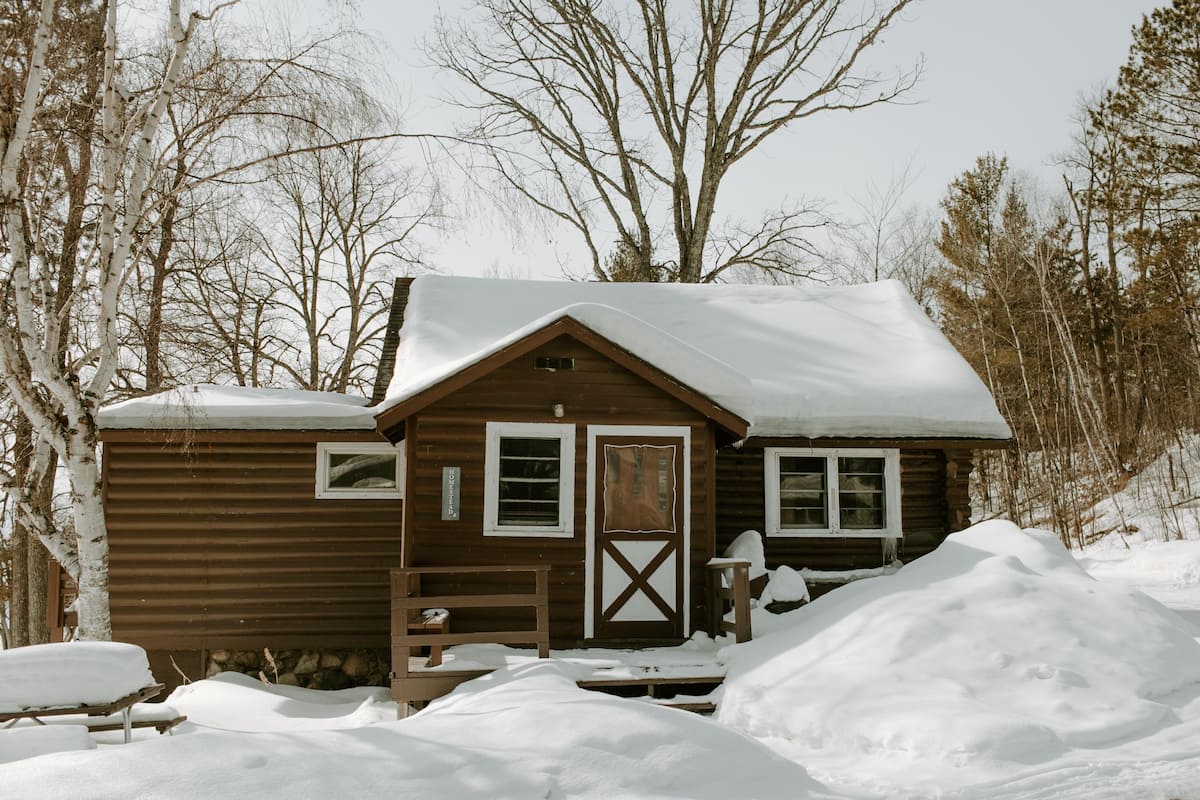 Una cabaña de madera con techo nevado, puerta de madera y ventanas.