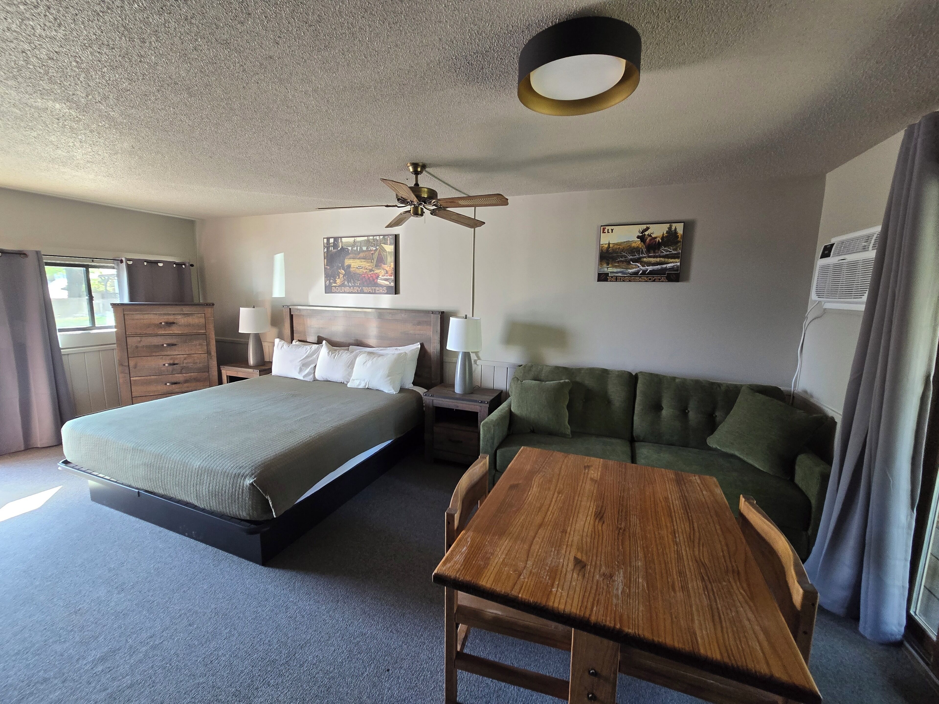 A hotel room with a bed, a green sofa, a wooden table, a ceiling fan, and a painting on the wall.