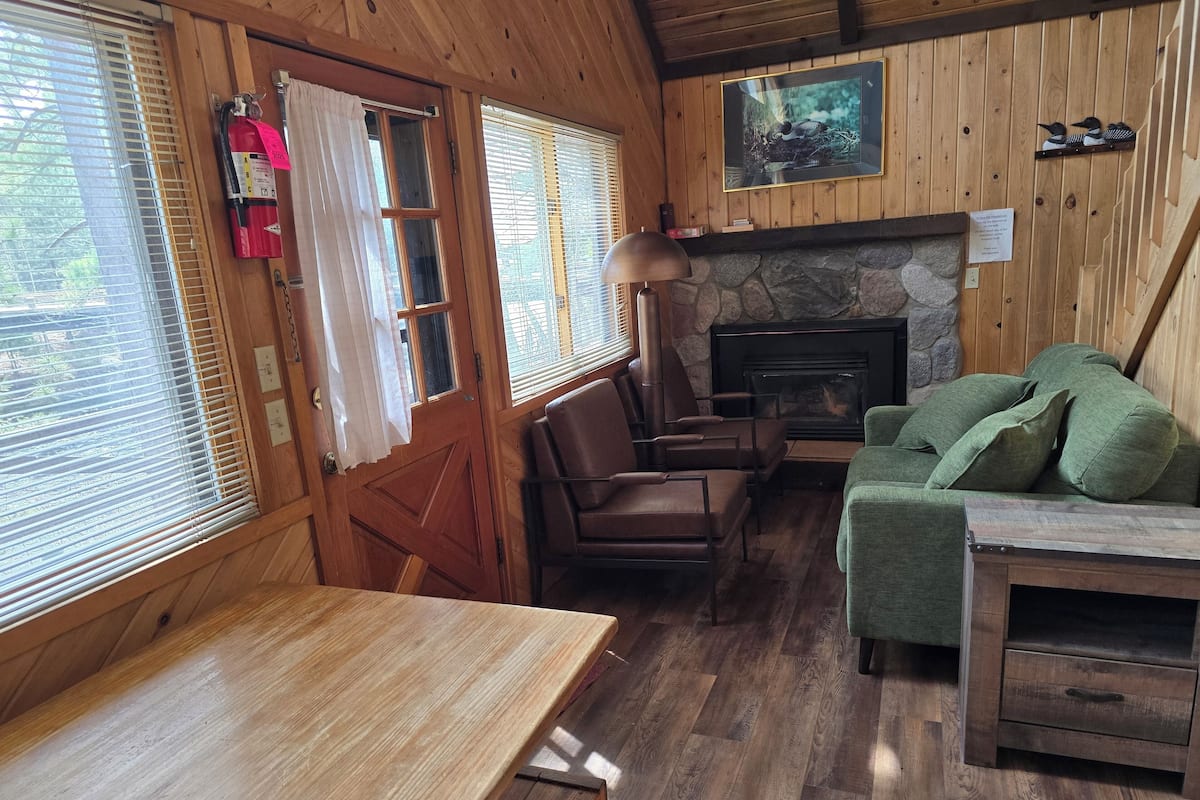 Interior acogedor de una cabaña con chimenea de piedra, paredes de madera, una mesa de comedor y asientos cómodos.