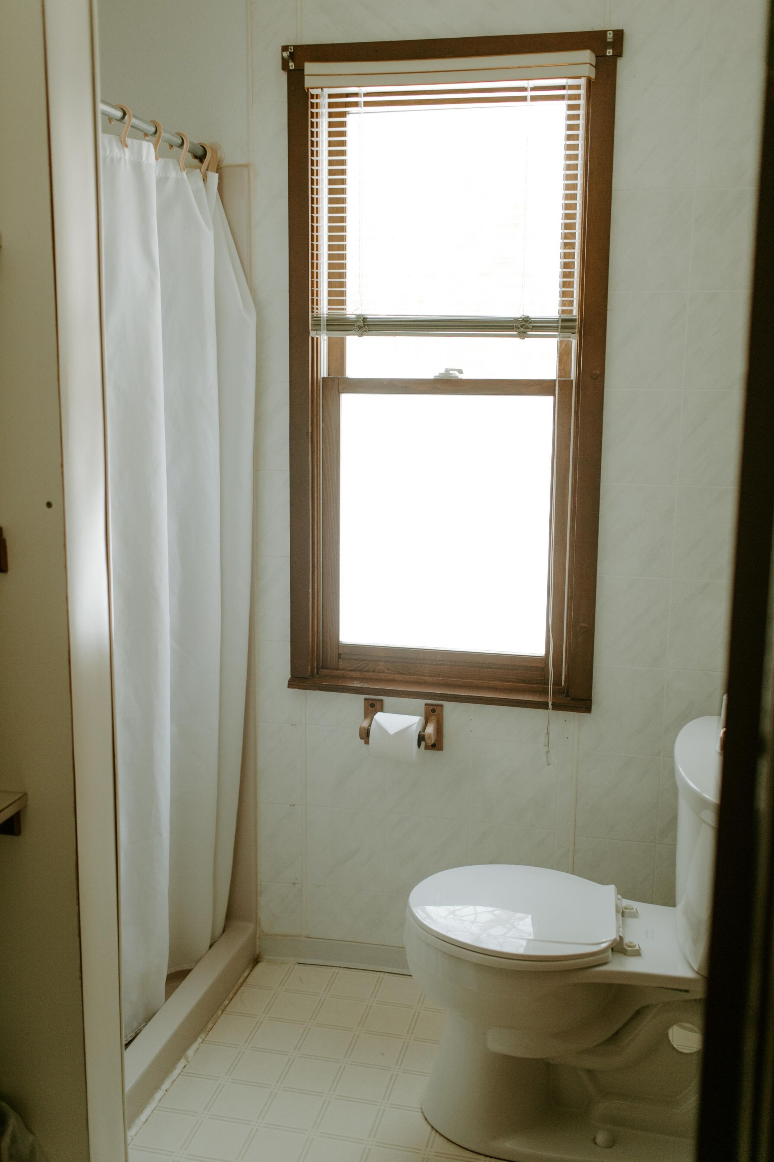 Un baño pequeño y sencillo con inodoro, ventana con persianas y cortina de ducha.