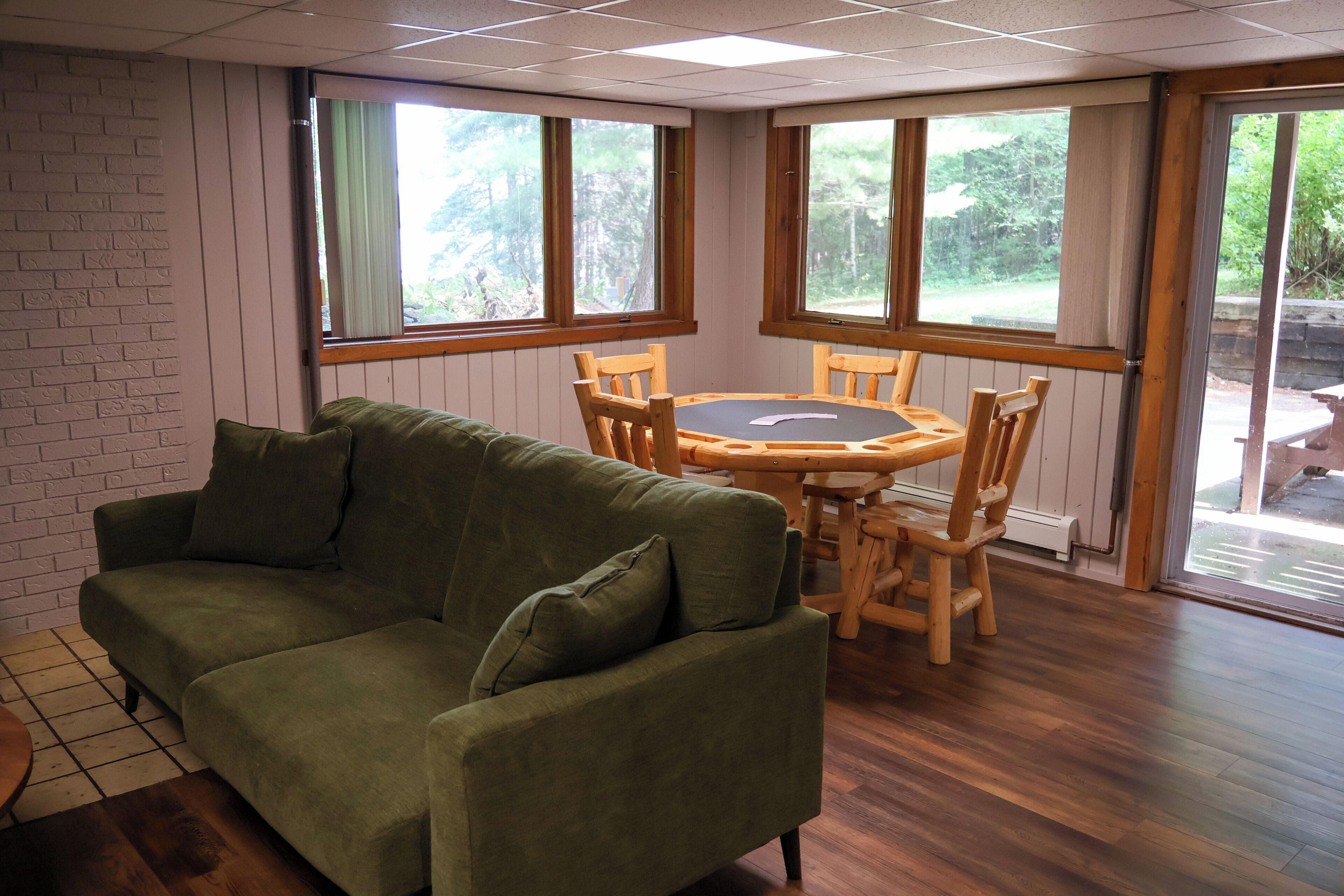 A room with a green sofa, wooden dining table, and chairs, large windows with curtains, and a brick wall.