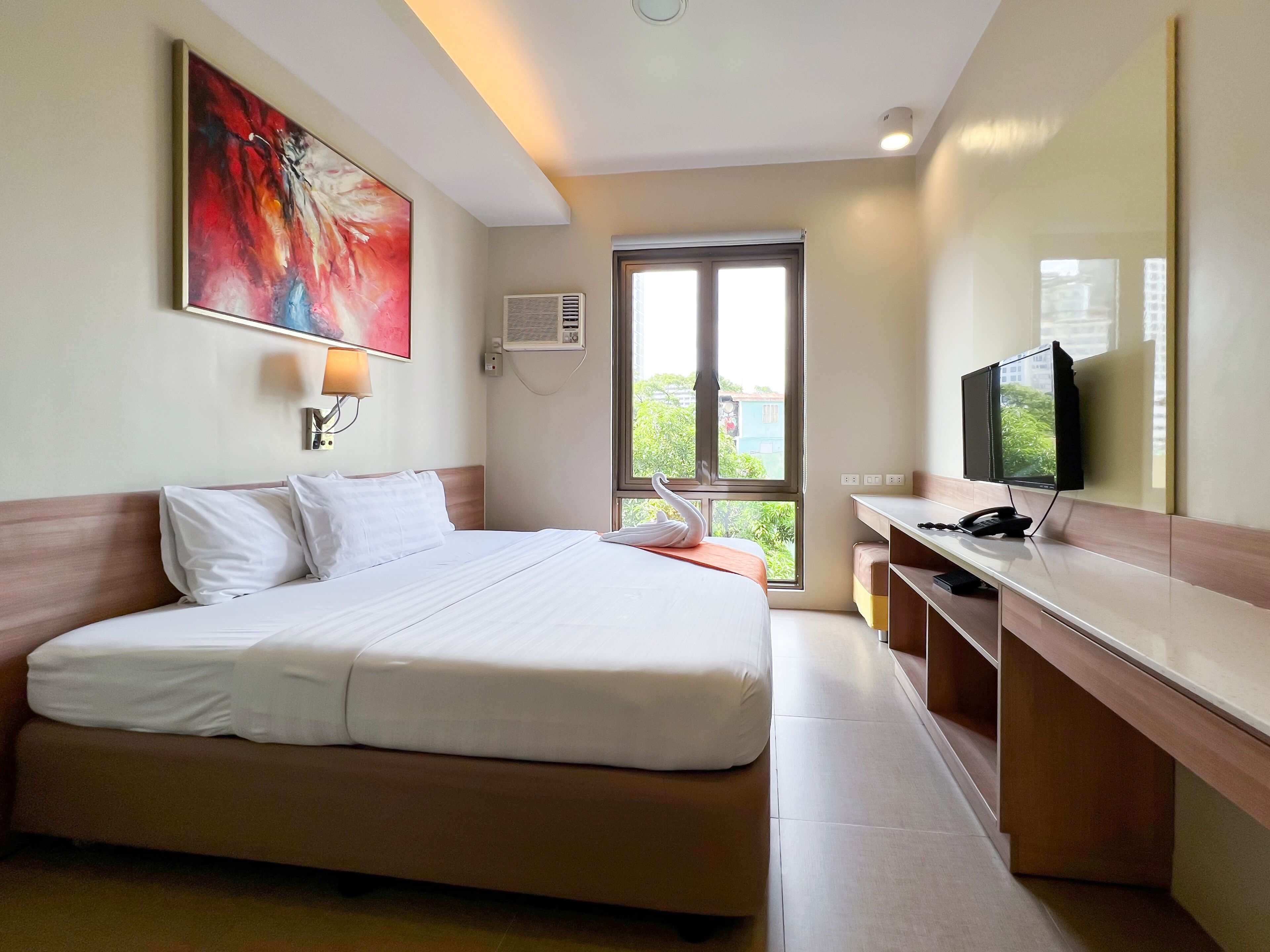 Una habitación de hotel con una cama grande, un escritorio con televisor de pantalla plana, una ventana con vista a follaje y un equipo de aire acondicionado.