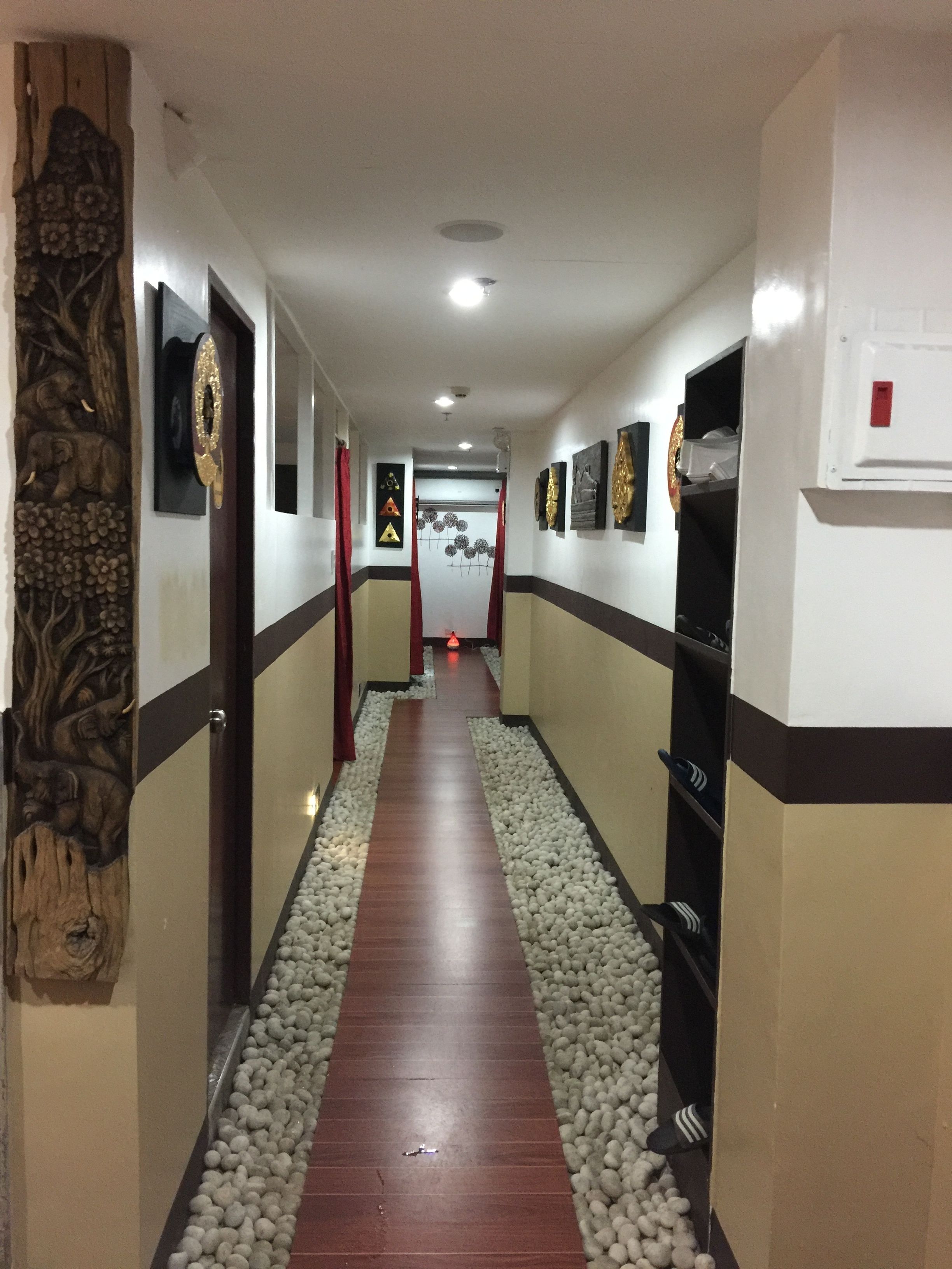 Un pasillo con piso de madera, paredes blancas y alfombra roja. Se observan cuadros enmarcados en las paredes y una puerta con panel decorativo a la izquierda.