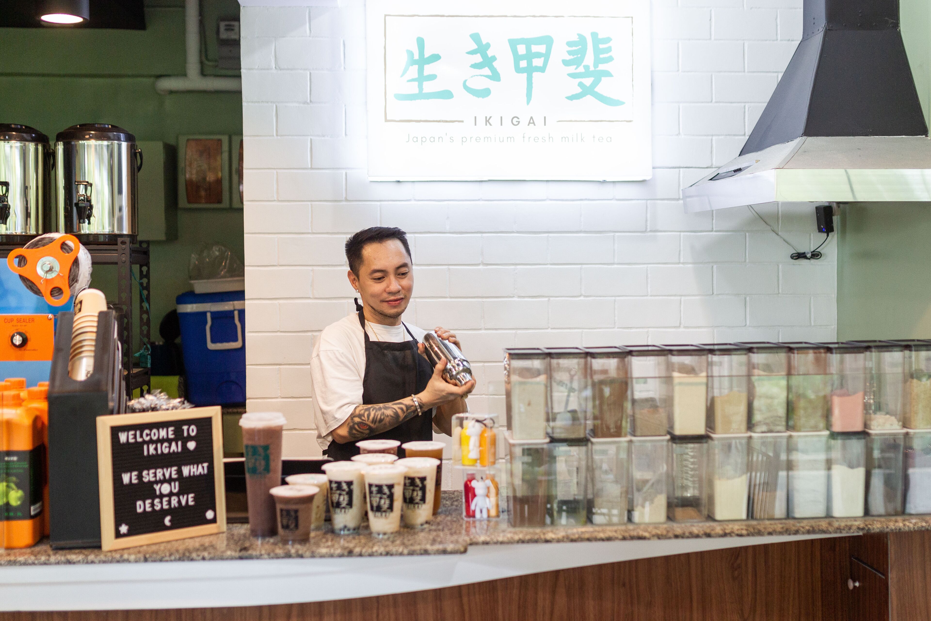 Ein Café-Interieur mit einer Theke, auf der verschiedene Kaffeebohnen ausgestellt sind, und einem Schild mit der Aufschrift 'Welcome to Ikigai'.