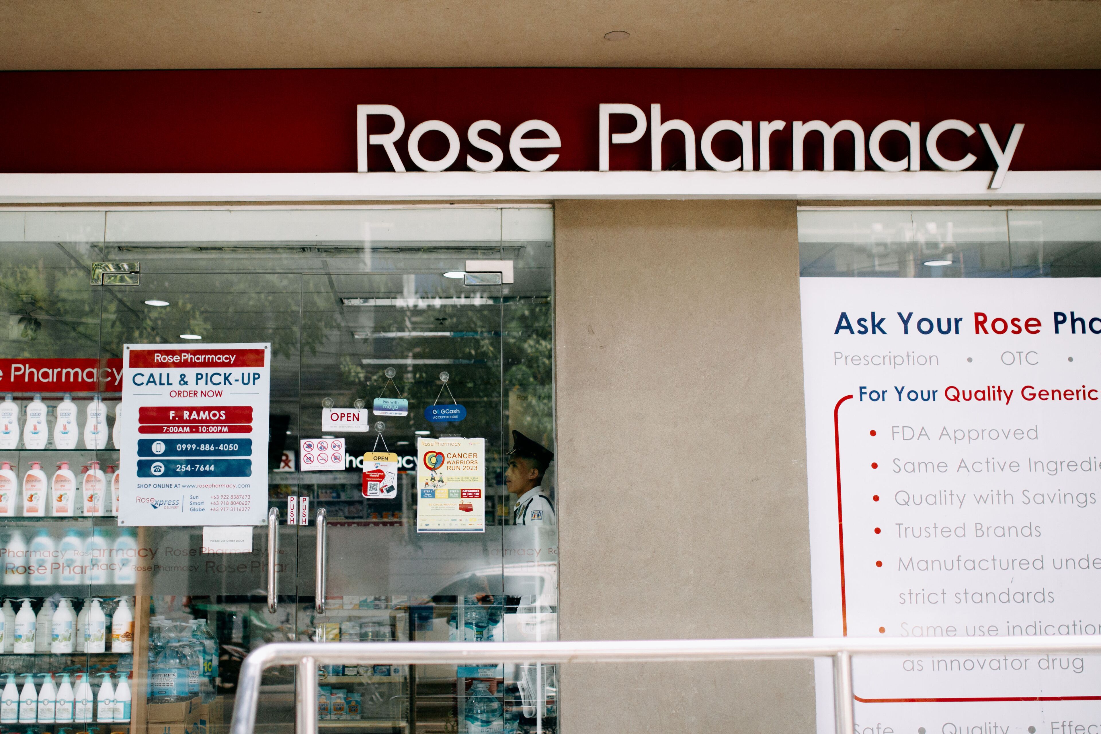 A pharmacy named Rose Pharmacy with a signboard advertising a call and pick-up service, a pharmacist inside, and various products on display.