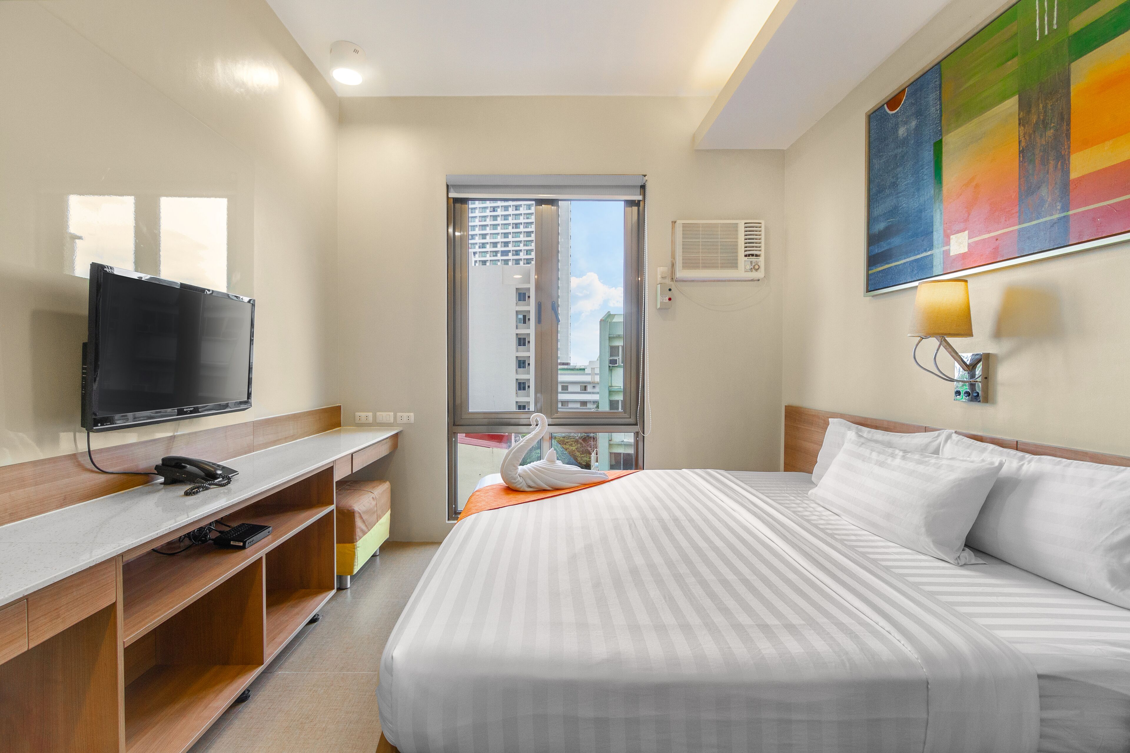 Habitació d'hotel amb un llit, una televisió, una taula de treball i una finestra que mostra edificis.