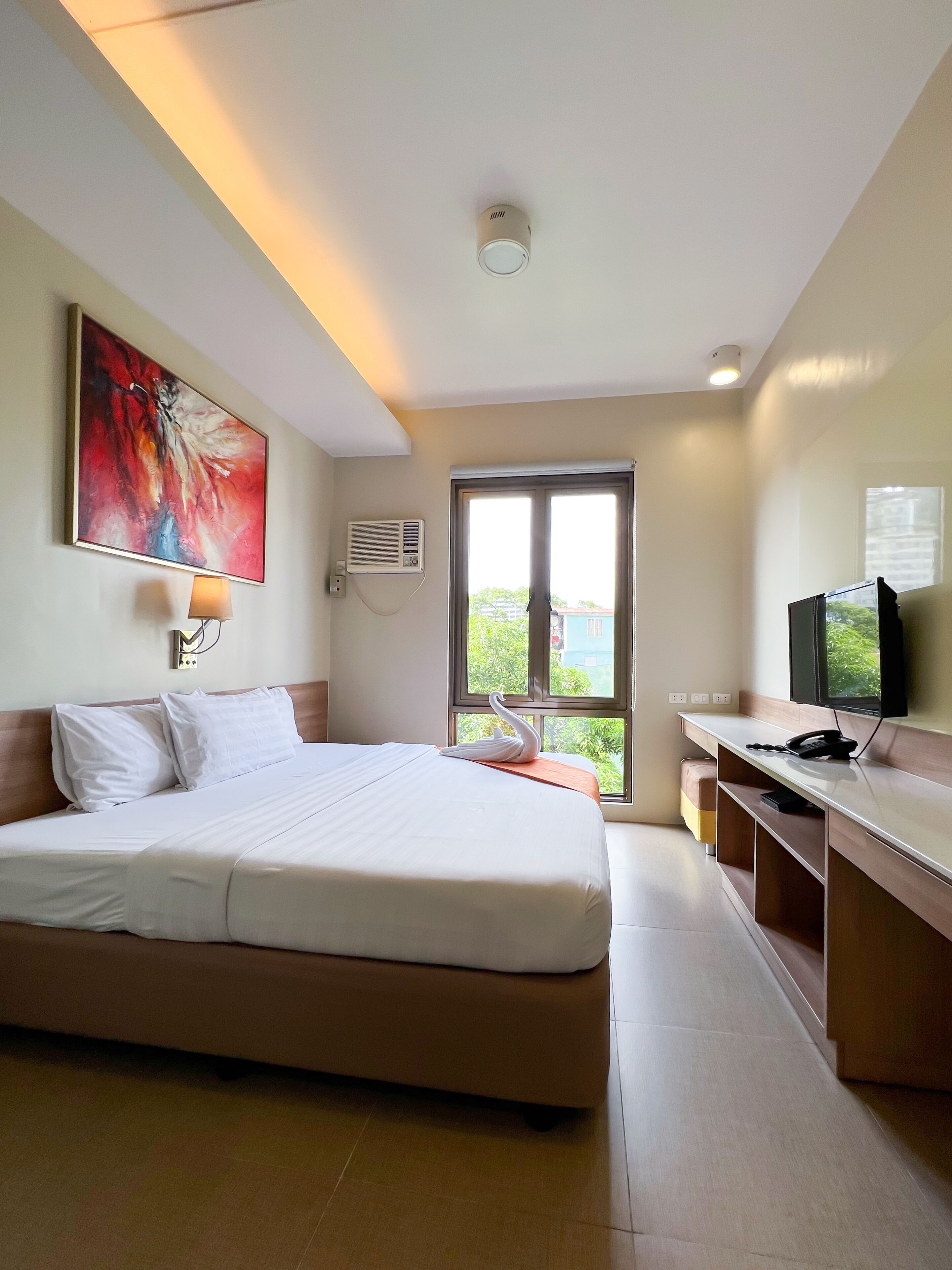 A hotel room with a large bed, a desk with a computer and phone, a TV, an air conditioning unit, and a window with a view of greenery.