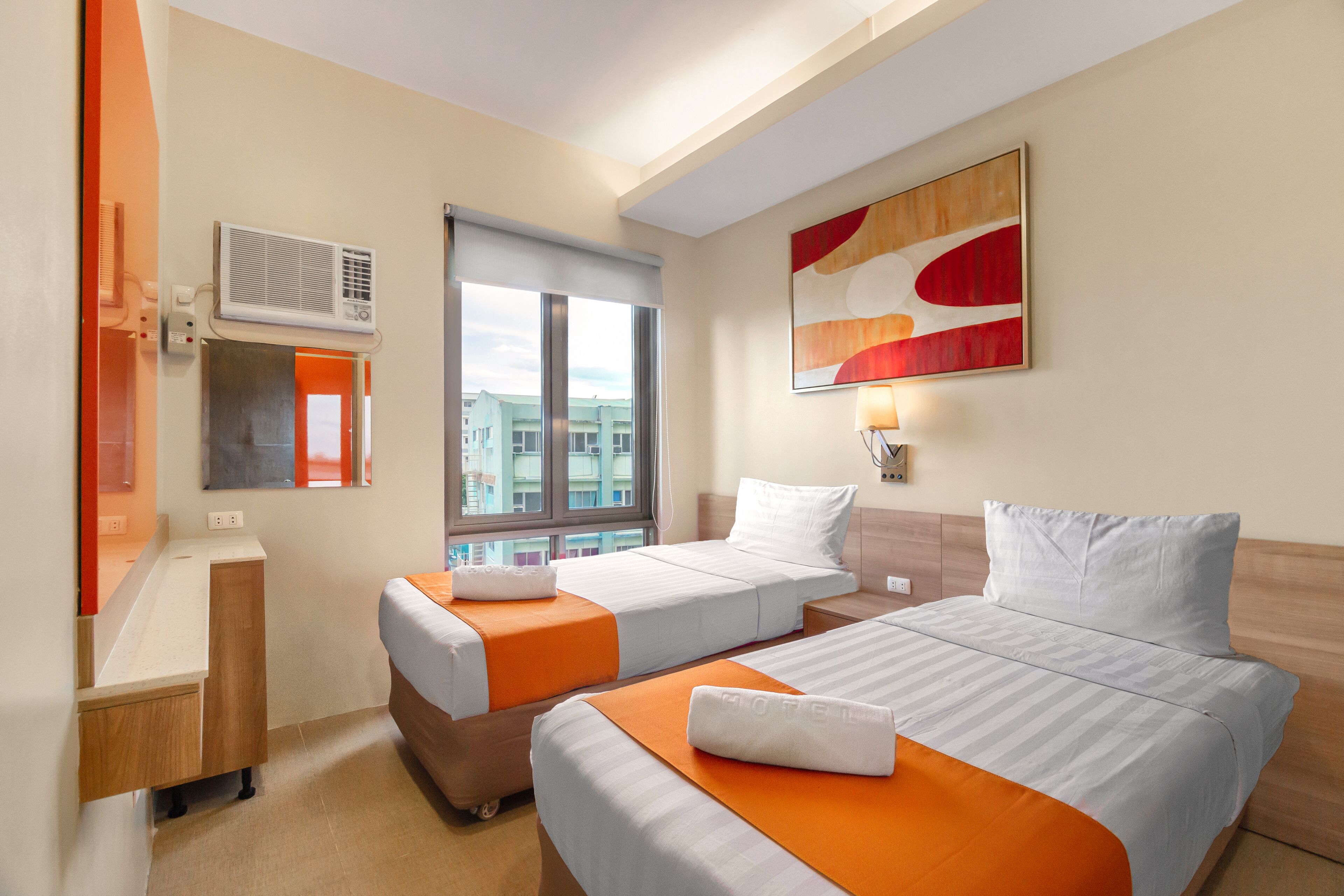A hotel room with two beds, a window with a view, an air conditioning unit, and a painting on the wall.