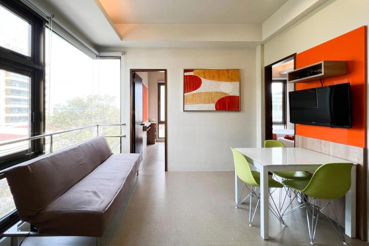 A modern hotel room with a sofa, a dining table with chairs, a flat-screen TV, and a large window.