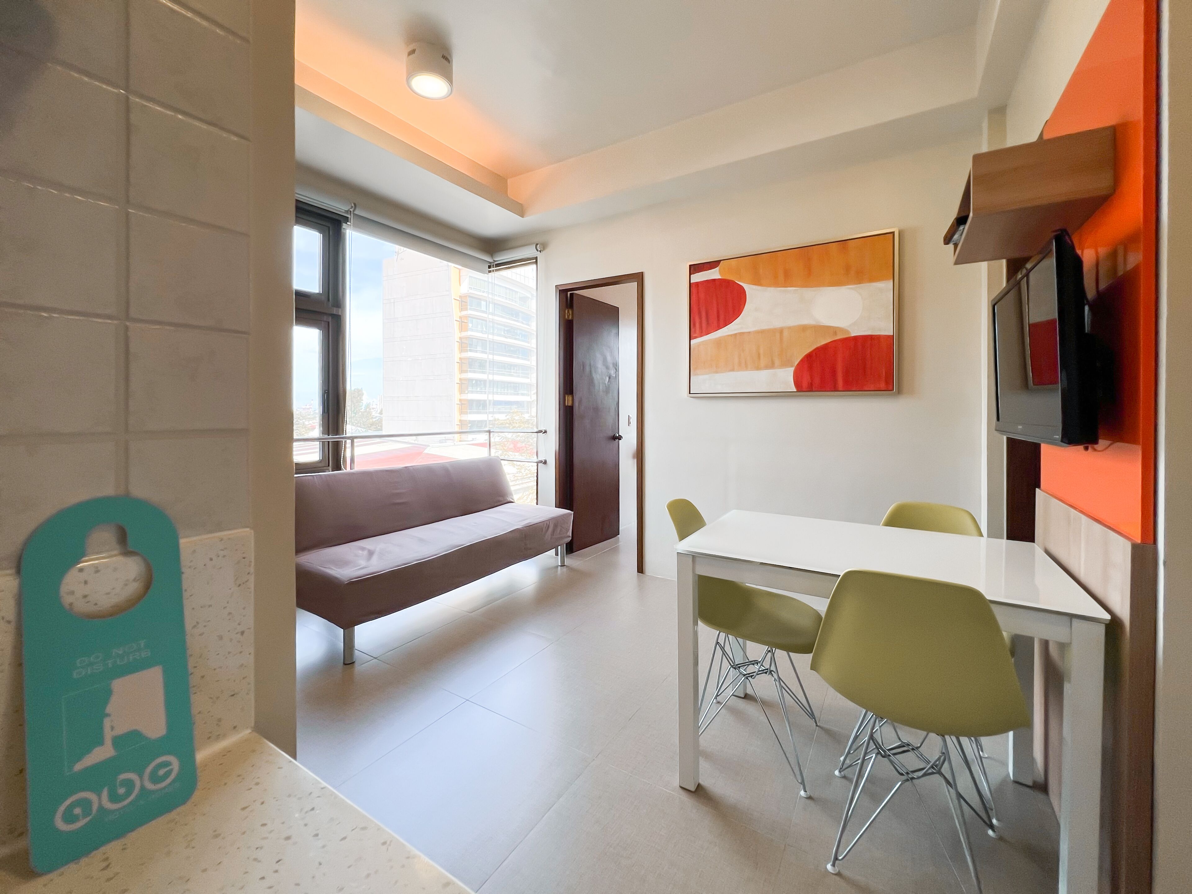 A modern living room with a sofa, a dining table with chairs, a TV mounted on the wall, and a window providing a view of the city.