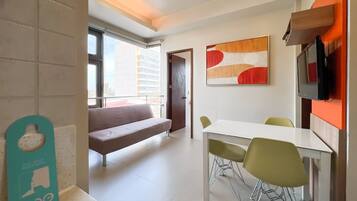 A modern living room with a sofa, a dining table with chairs, a TV mounted on the wall, and a window providing a view of the city.