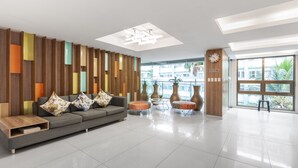 Un vestíbulo moderno con un sofá gris, paneles verticales coloridos, un mostrador de recepción de madera y un gran ventanal con vistas a palmeras.