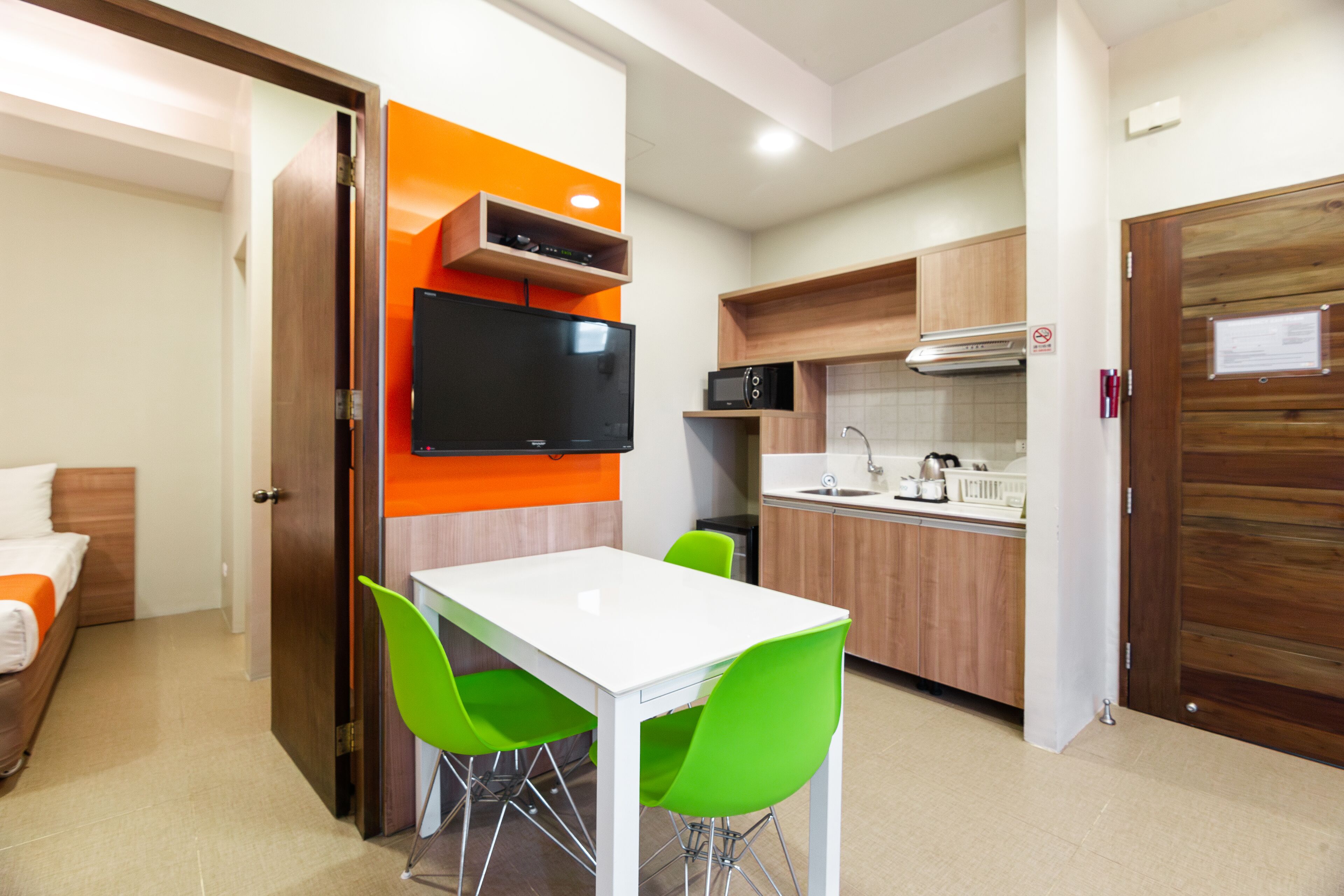 Quarto de hotel compacto com cozinha americana, área de jantar com mesa branca e cadeiras verdes, e cama com roupa de cama laranja.