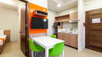 Quarto de hotel compacto com cozinha americana, área de jantar com mesa branca e cadeiras verdes, e cama com roupa de cama laranja.