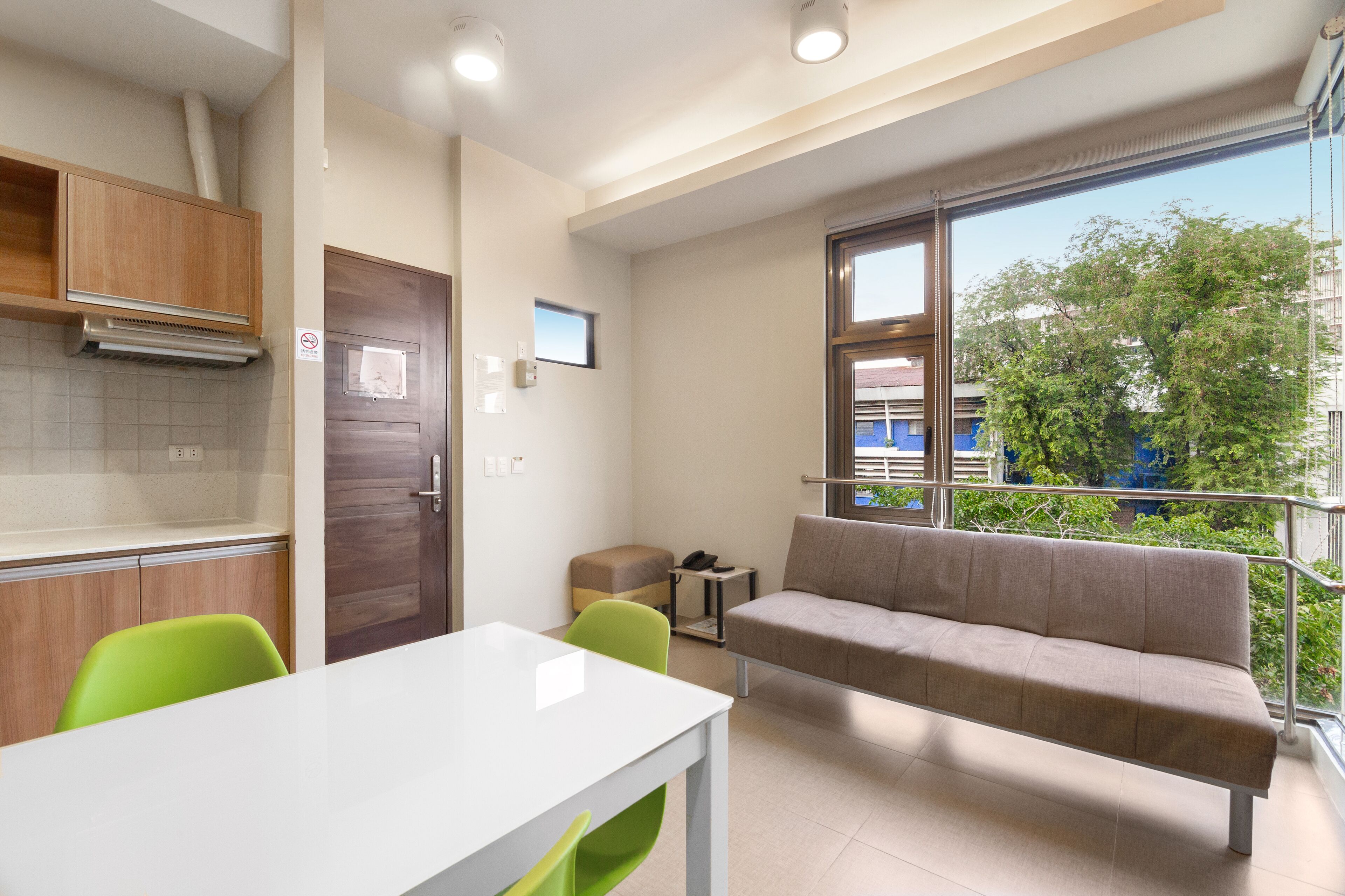 Un appartement moderne doté d’une kitchenette, d’un coin repas avec une table blanche et des chaises vertes, d’un canapé et d’un balcon offrant une vue sur les arbres et les immeubles.