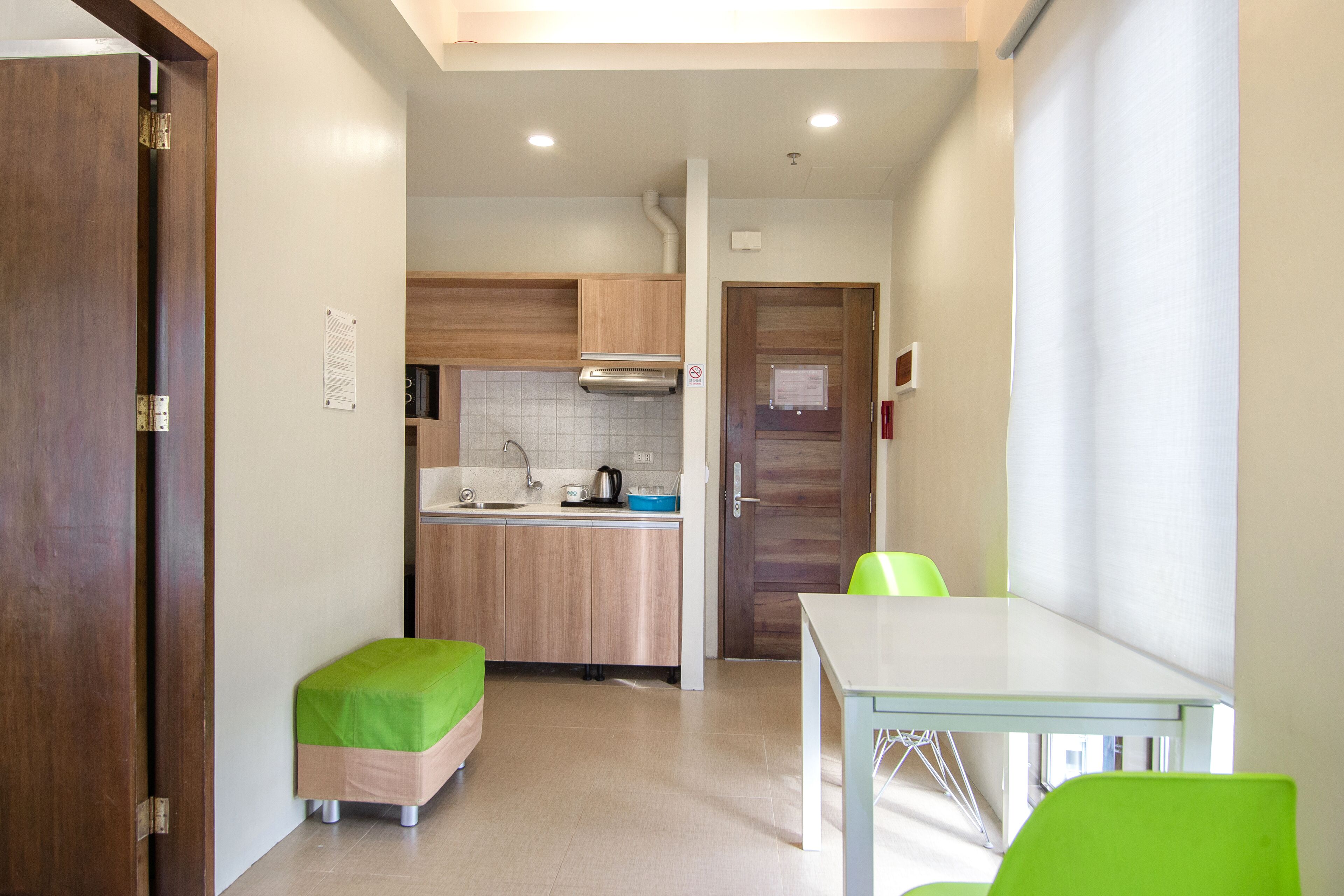 A compact kitchen with wooden cabinets, a sink, and a stove. There is a small table with a green chair, and a wooden door on the left.
