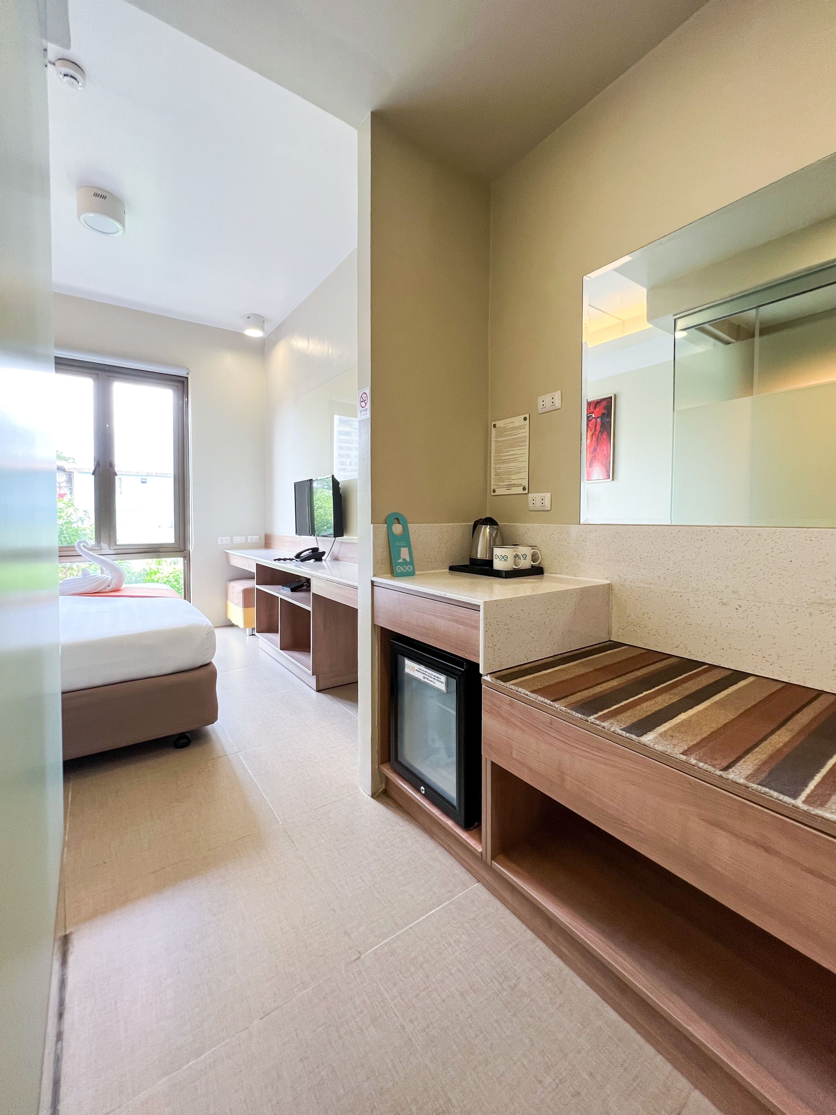 A modern hotel room with a bed, a small refrigerator, a coffee machine, and a mirror on the wall.