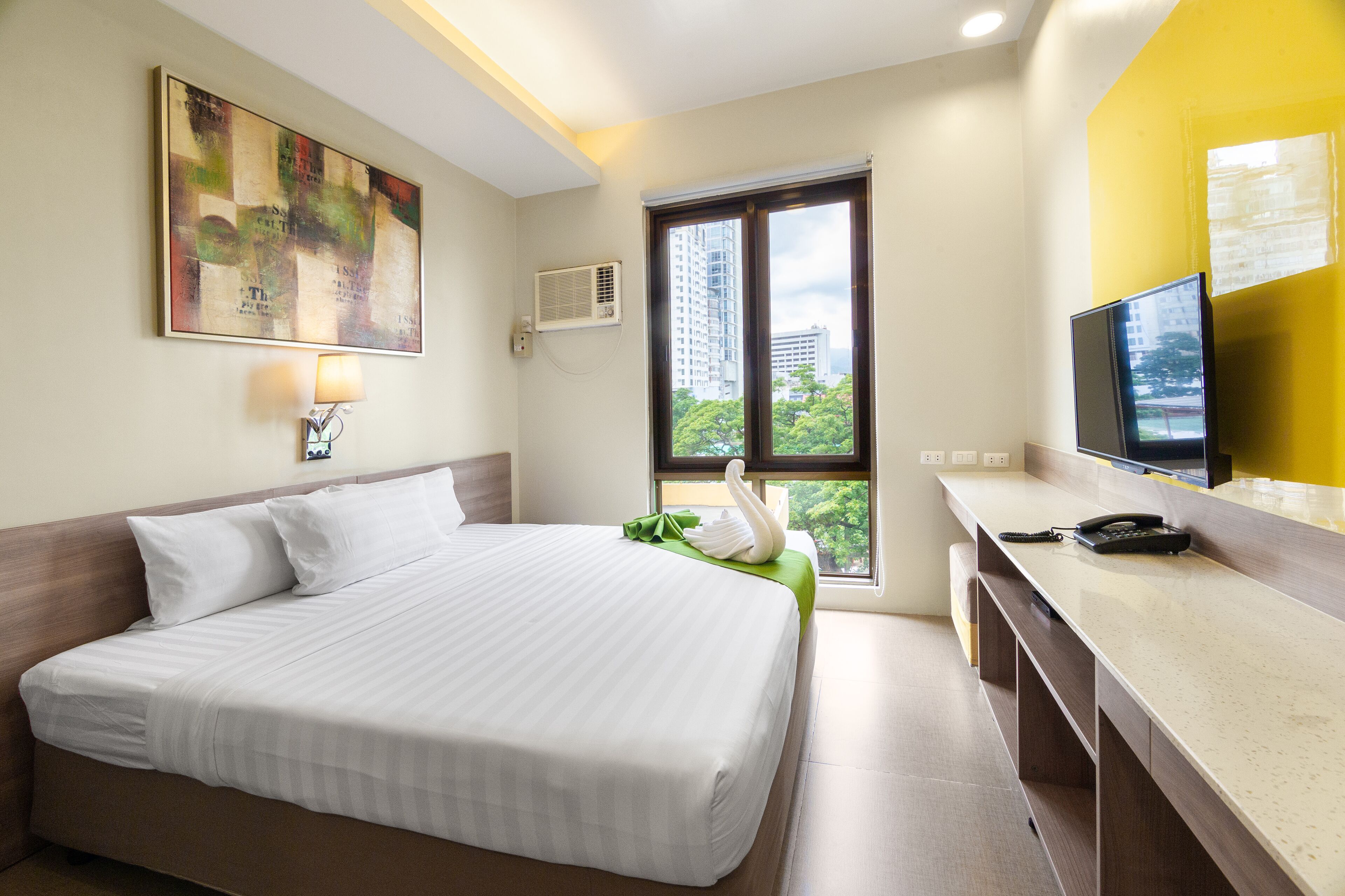 Habitación de hotel con cama, escritorio, teléfono, televisor y ventana con vista a edificios y árboles.