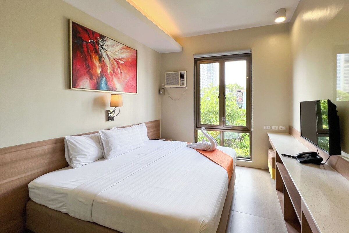 A hotel room with a bed, a desk with a telephone, a TV, a window with a view, and an air conditioning unit.