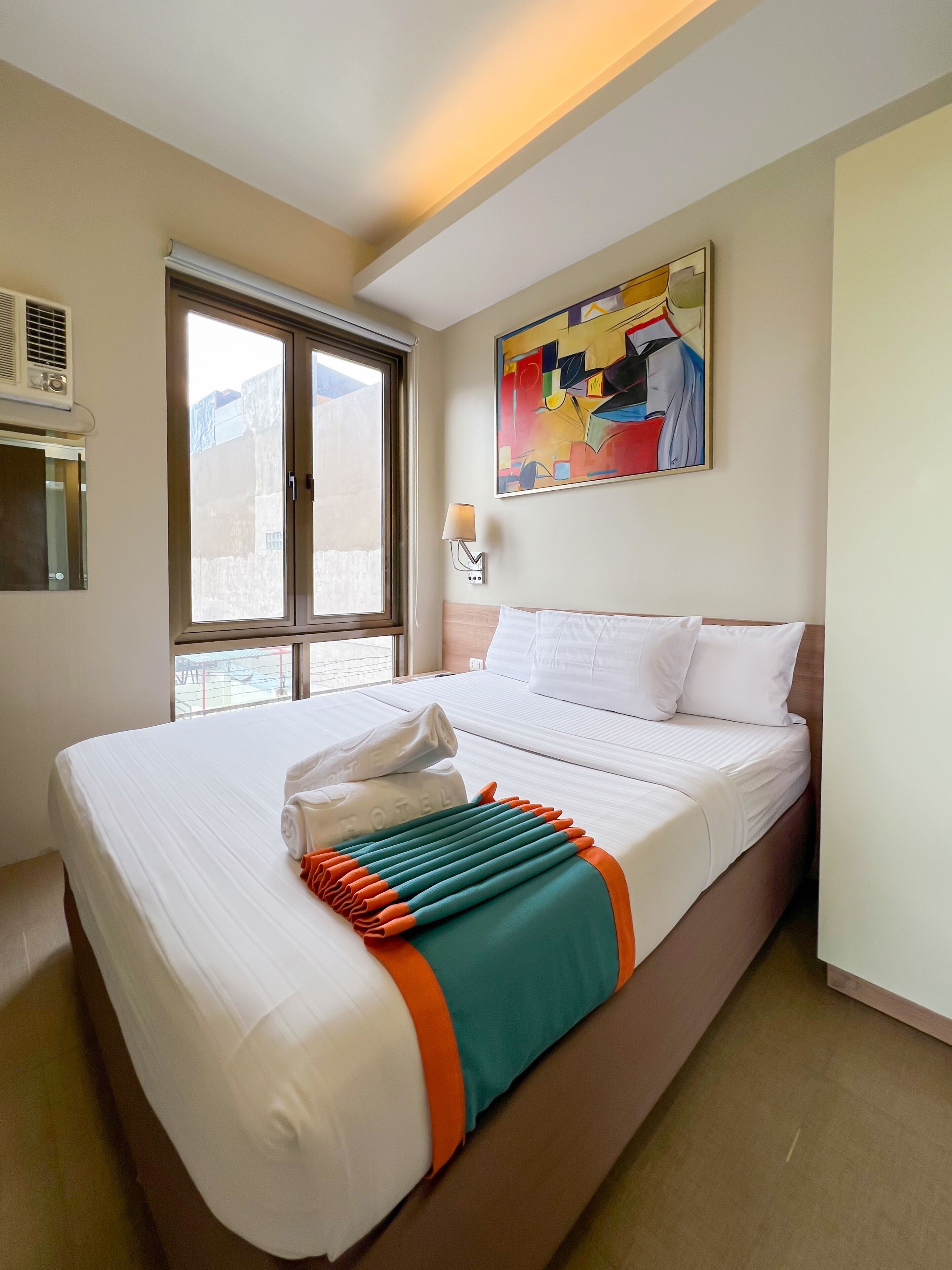 A hotel room with a large bed, a window with a view, a painting on the wall, and a lamp.