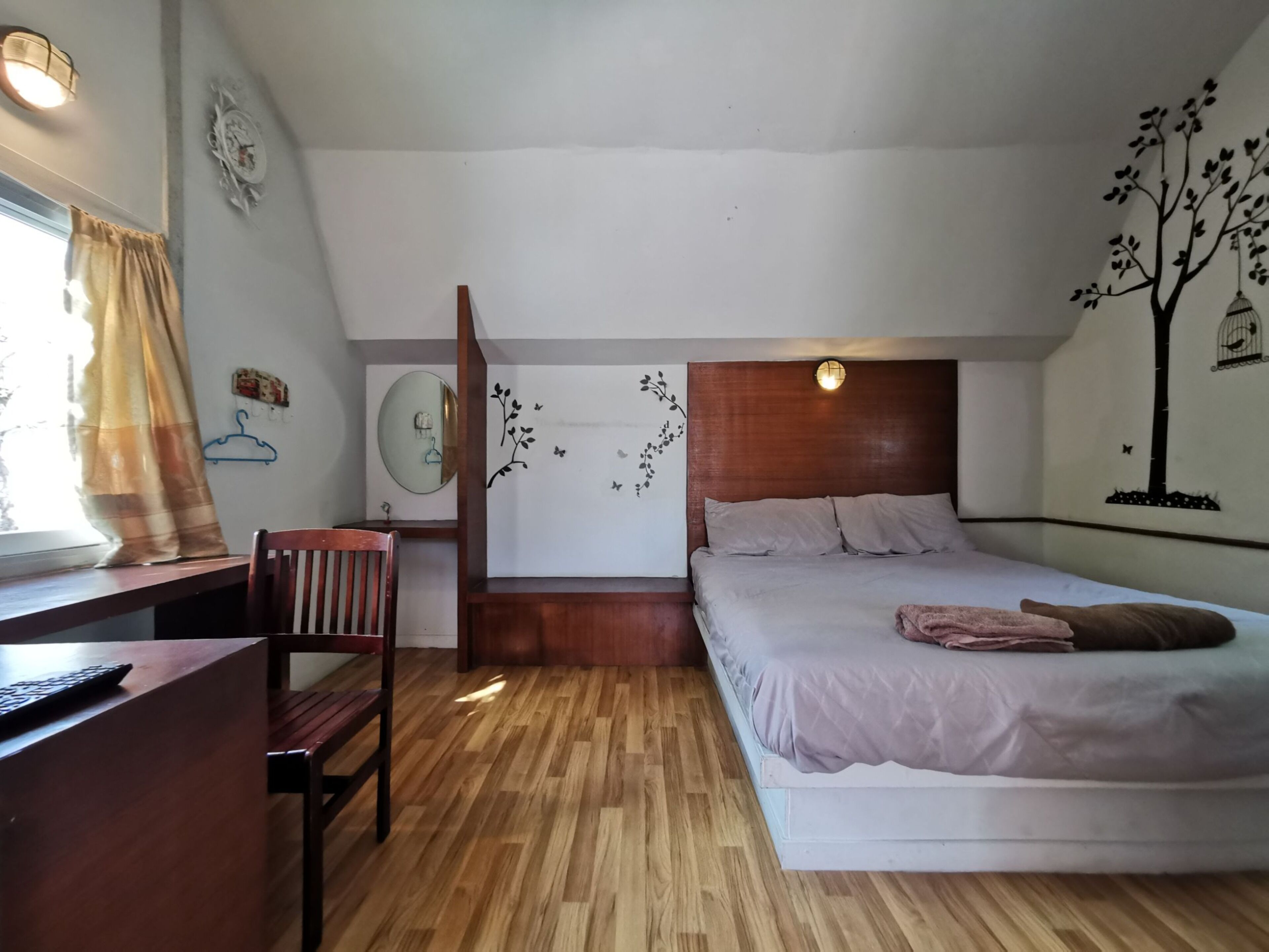 A bedroom with a wooden desk, a bed with a headboard, a mirror, and wall decorations including a tree and butterflies.