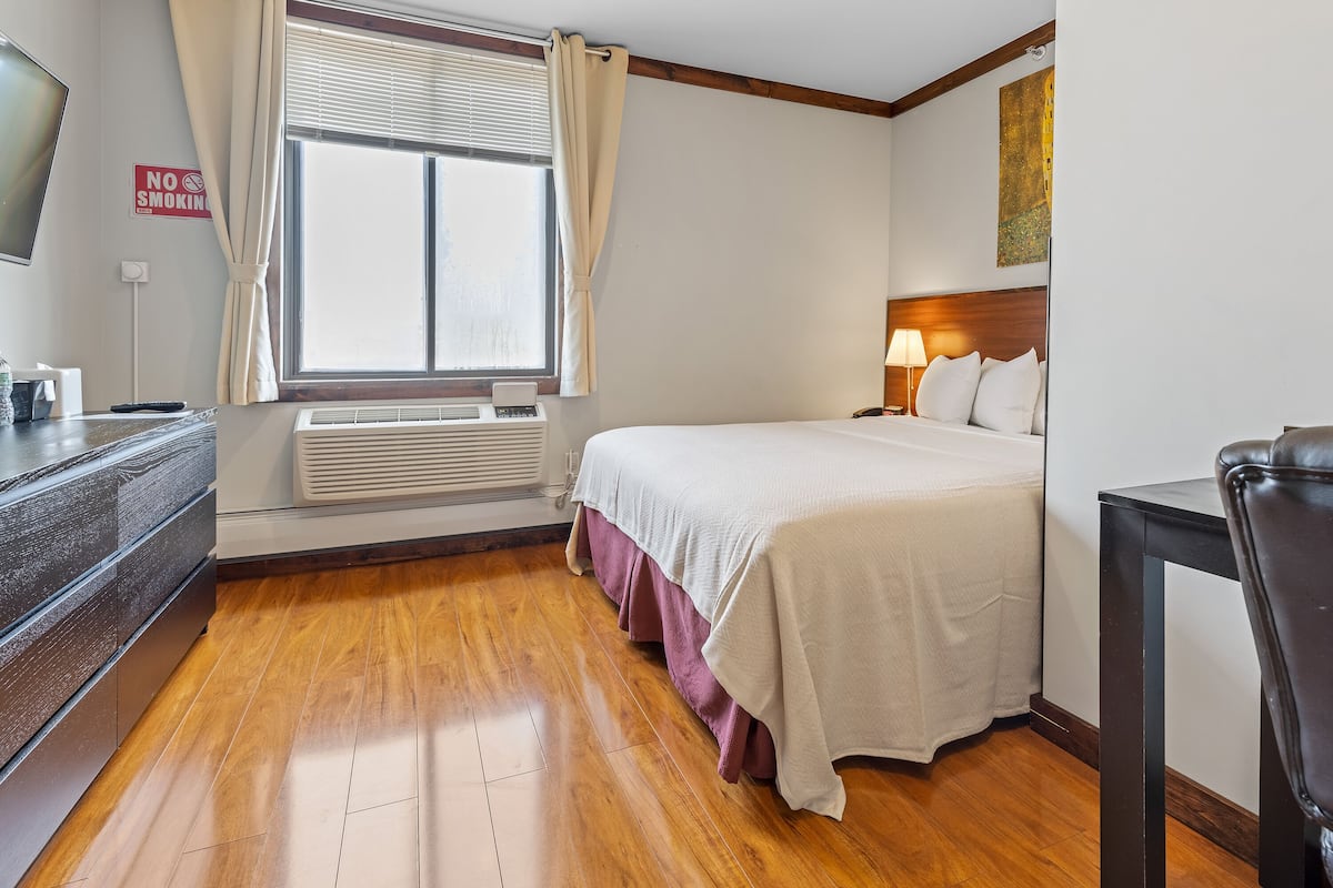 A hotel room with a bed, a desk, a chair, a window with curtains, a wall-mounted TV, and a 'No Smoking' sign.