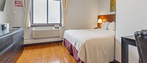 Habitación de hotel con cama, escritorio, silla, ventana con cortinas, televisor montado en la pared y un cartel de 'No Fumar'.