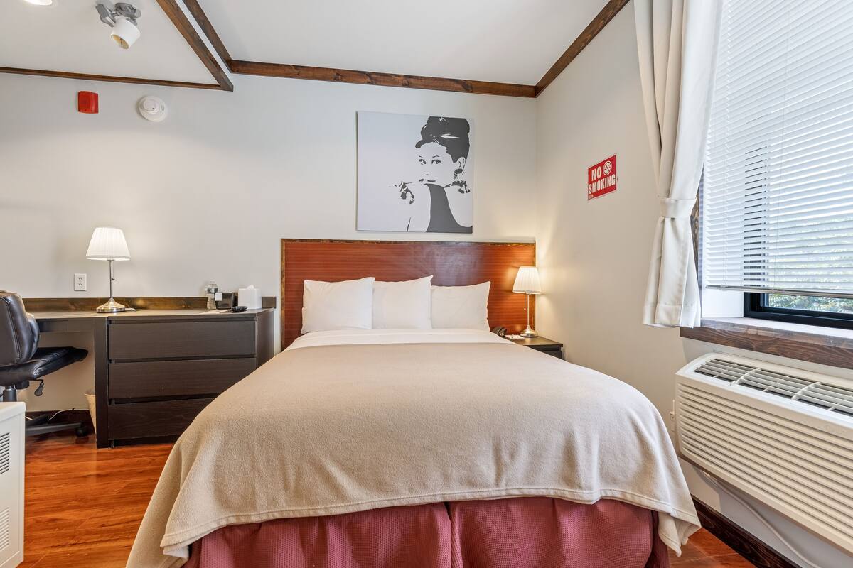 A hotel room with a bed, bedside tables, a desk, a chair, a lamp, a wall art piece, a window with blinds, and a no smoking sign.