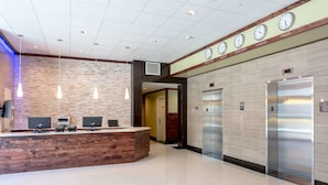 Un hall d’hôtel avec un comptoir d’accueil en bois, plusieurs horloges indiquant l’heure de différentes villes et deux ascenseurs.