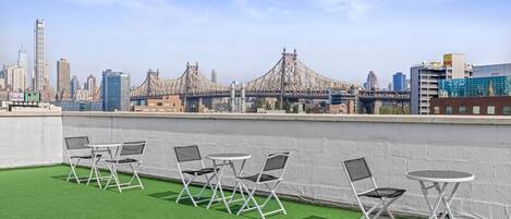Une terrasse sur le toit, avec un revêtement d’herbe artificielle, des chaises en métal blanc et une petite table ronde. On aperçoit, en arrière-plan, le profil de la ville et un pont.