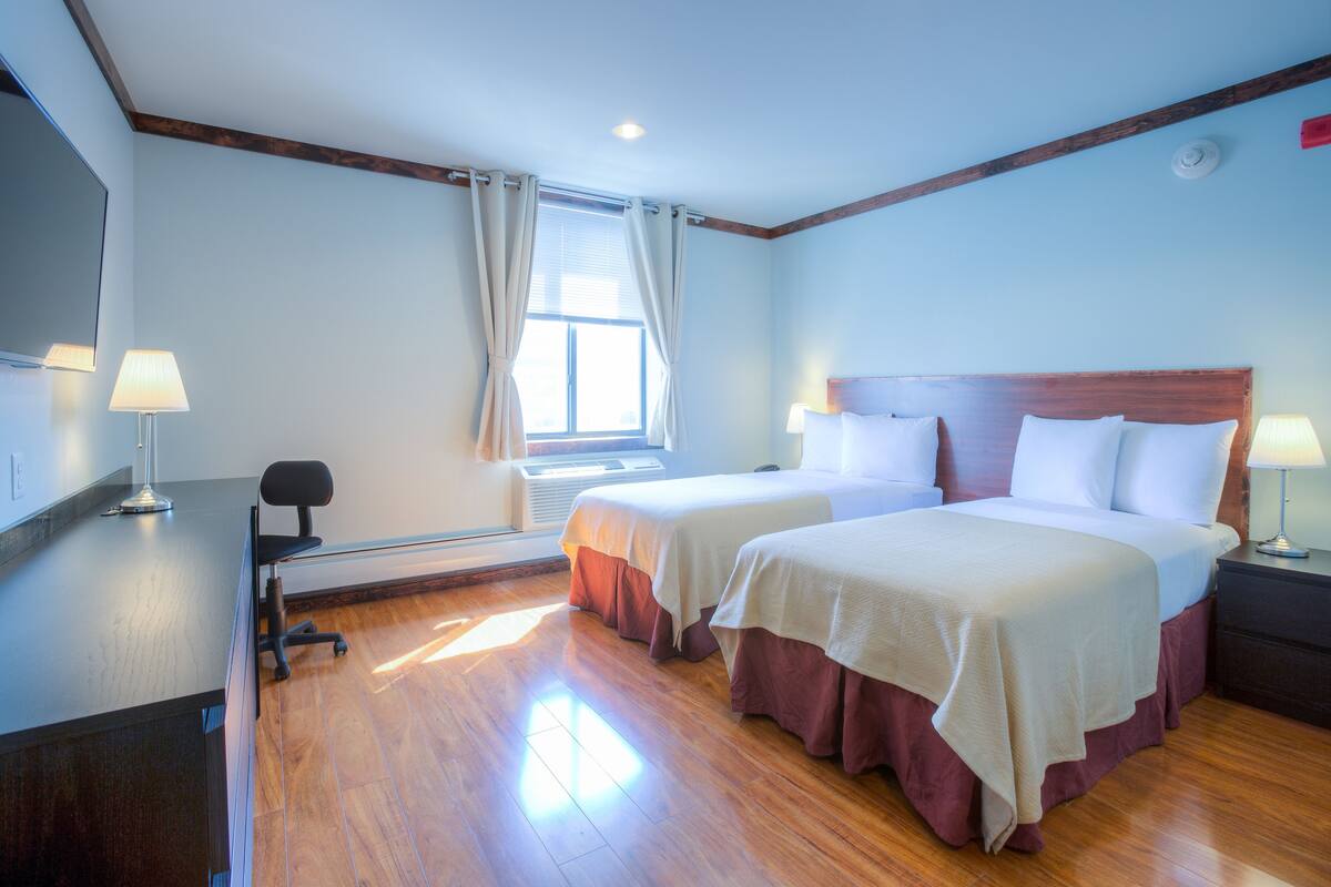 A hotel room with two beds, wooden flooring, a desk, and a window with curtains.