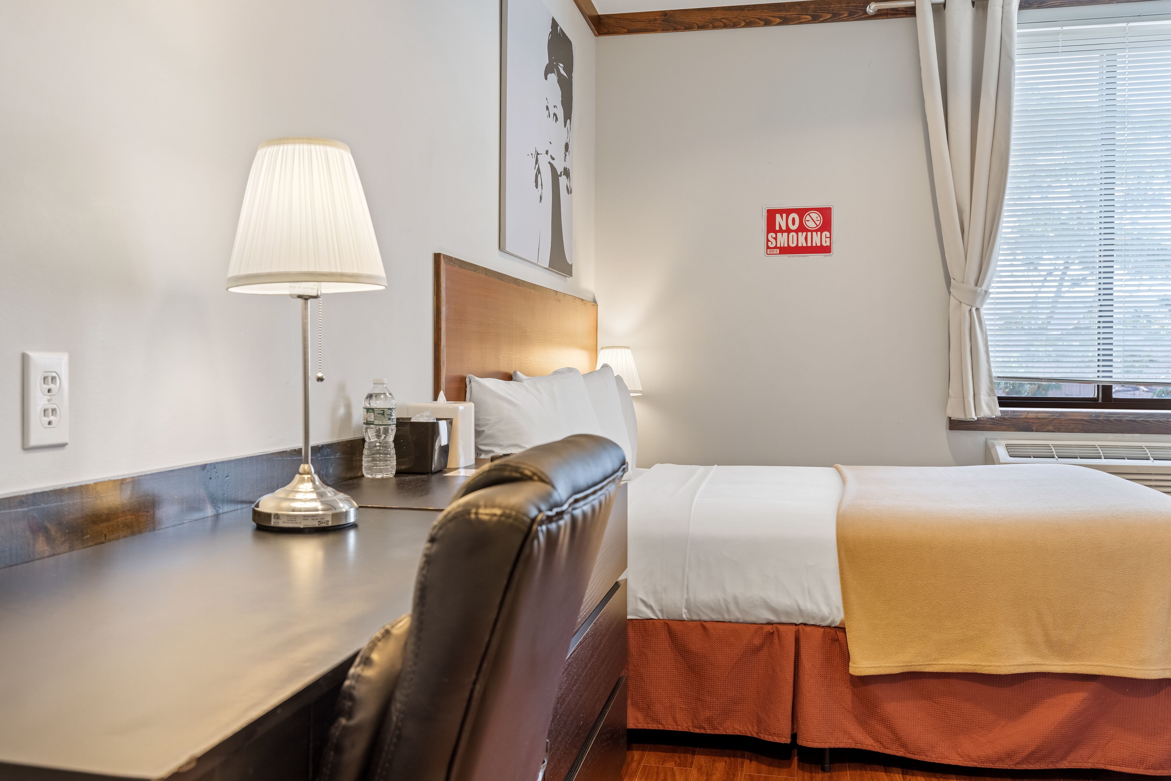 Een hotelkamer met een bed, een bureau met lamp, een bordje ‘Niet roken’ en een raam met gordijnen.