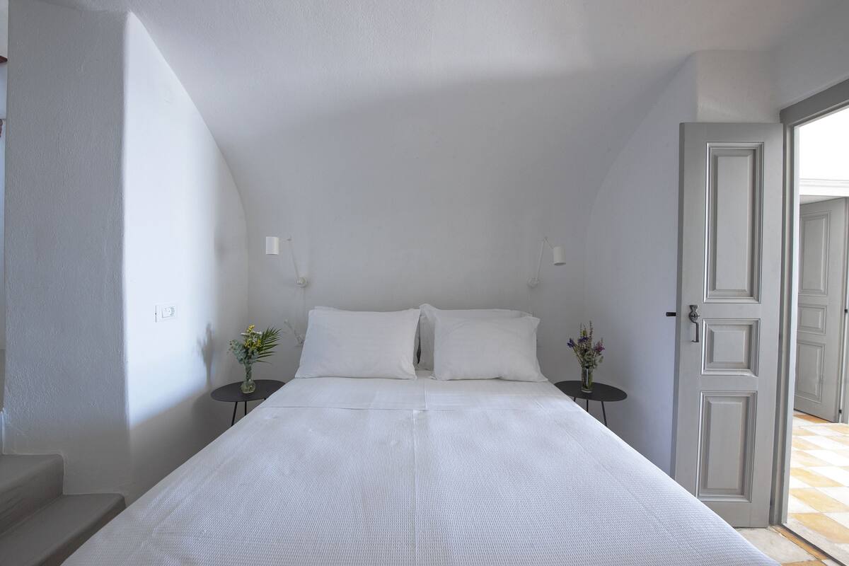 Un dormitorio con una cama grande, dos mesitas de noche con lámparas y una puerta que da a otra habitación.