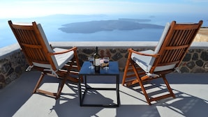 Two wooden chairs with cushions, a small table, and a bottle of wine on a balcony overlooking a scenic view. - Ikia Kriton (Santorini)