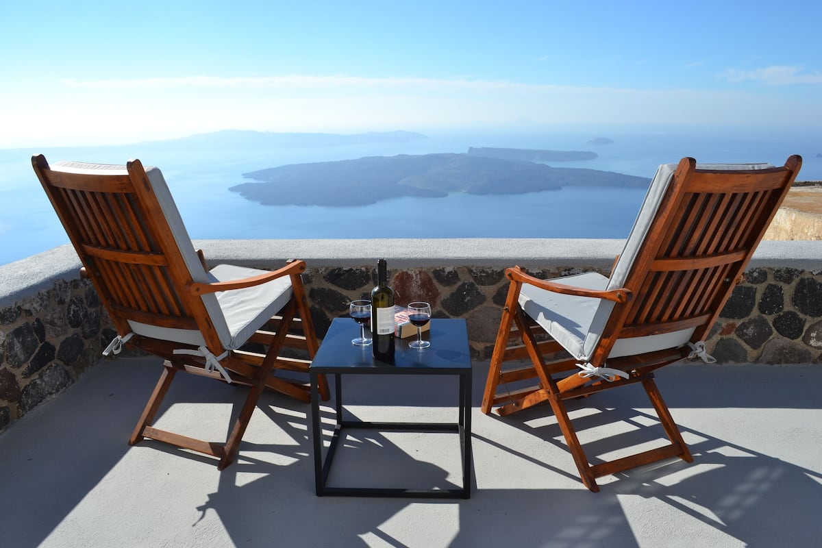 Dos sillas de madera con cojines, una mesita y una botella de vino en un balcón con vistas panorámicas.