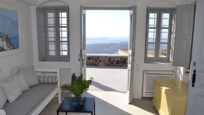 A room with a white sofa, a small table with a vase, and a view of the sea through open doors. - Ikia Kriton (Santorini)